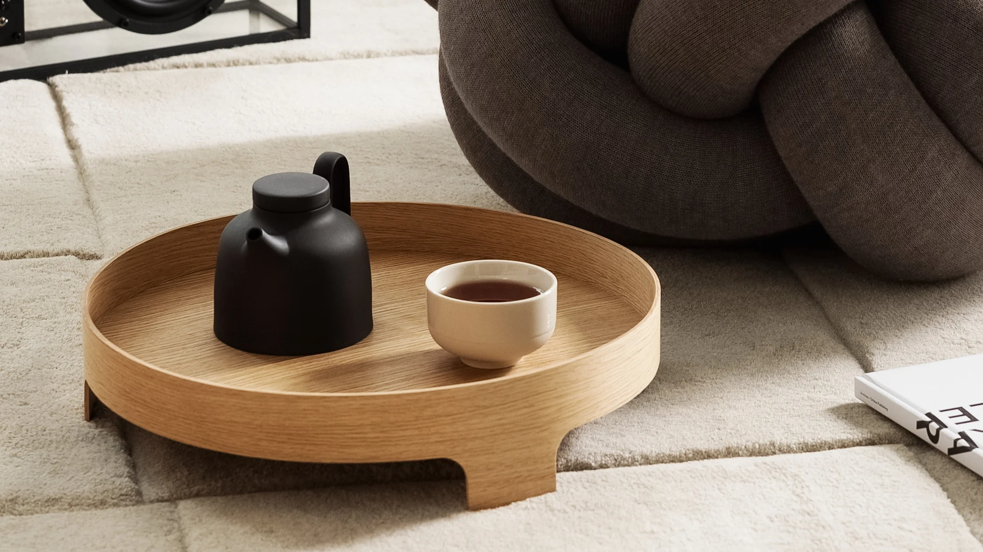 Lagom : Le plateau Bridge est posé sur un tapis, sur lequel se trouvent une théière et une tasse de thé, avec en arrière-plan le coussin XL, le tout de Design House Stockholm.