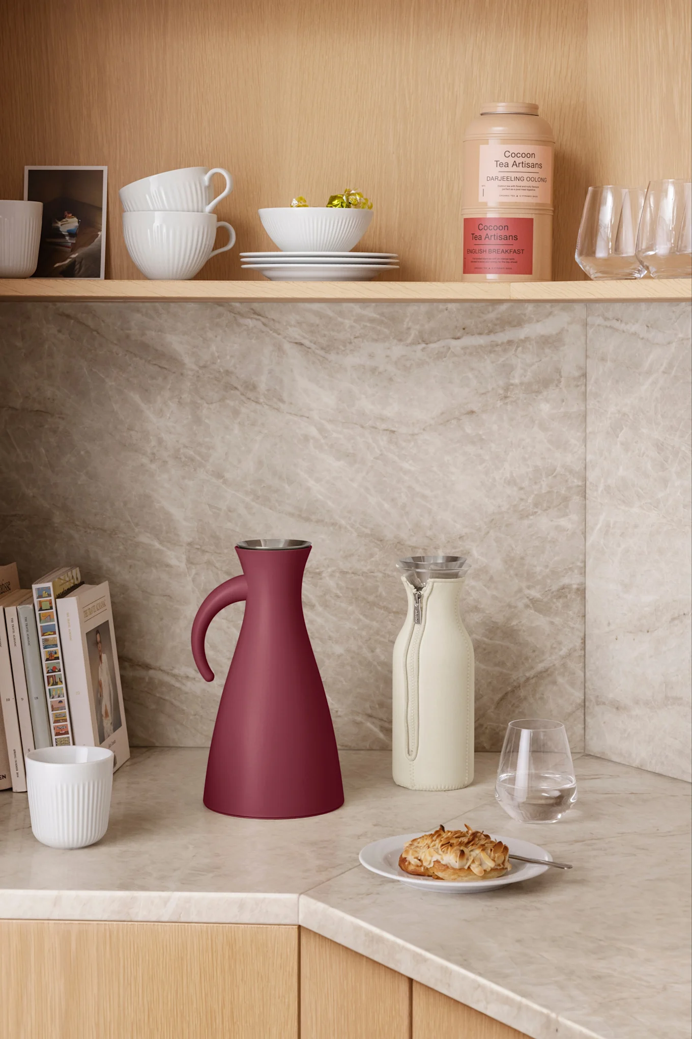 Une bouteille thermos conique rouge foncé et un pichet crème clair posés sur un plan de travail en marbre. Des bols, des tasses et des boîtes à thé blancs sont disposés sur les étagères au-dessus.