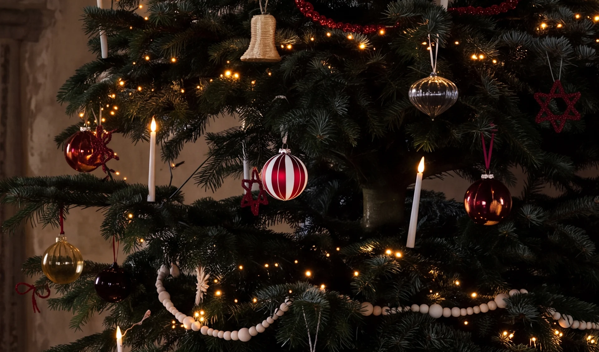 Un sapin de Noël décoré avec des lumières chaudes, des boules rouges, argentées et dorées, des bougies blanches et des guirlandes de perles en bois.