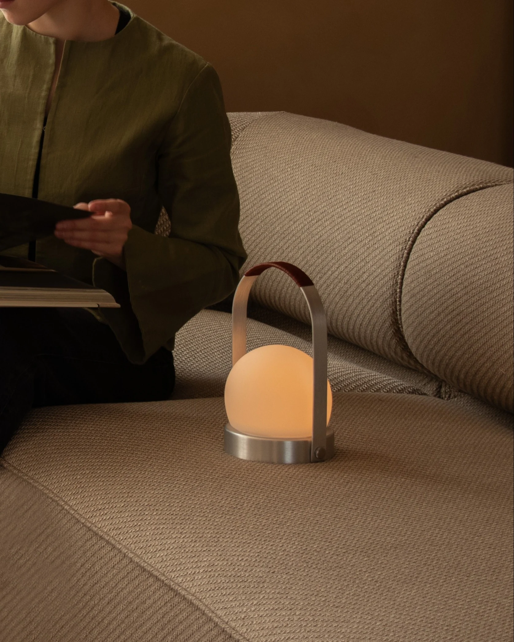 Une personne lit un livre sur un canapé beige à côté d'une lampe portable sphérique lumineuse avec une poignée en cuir.