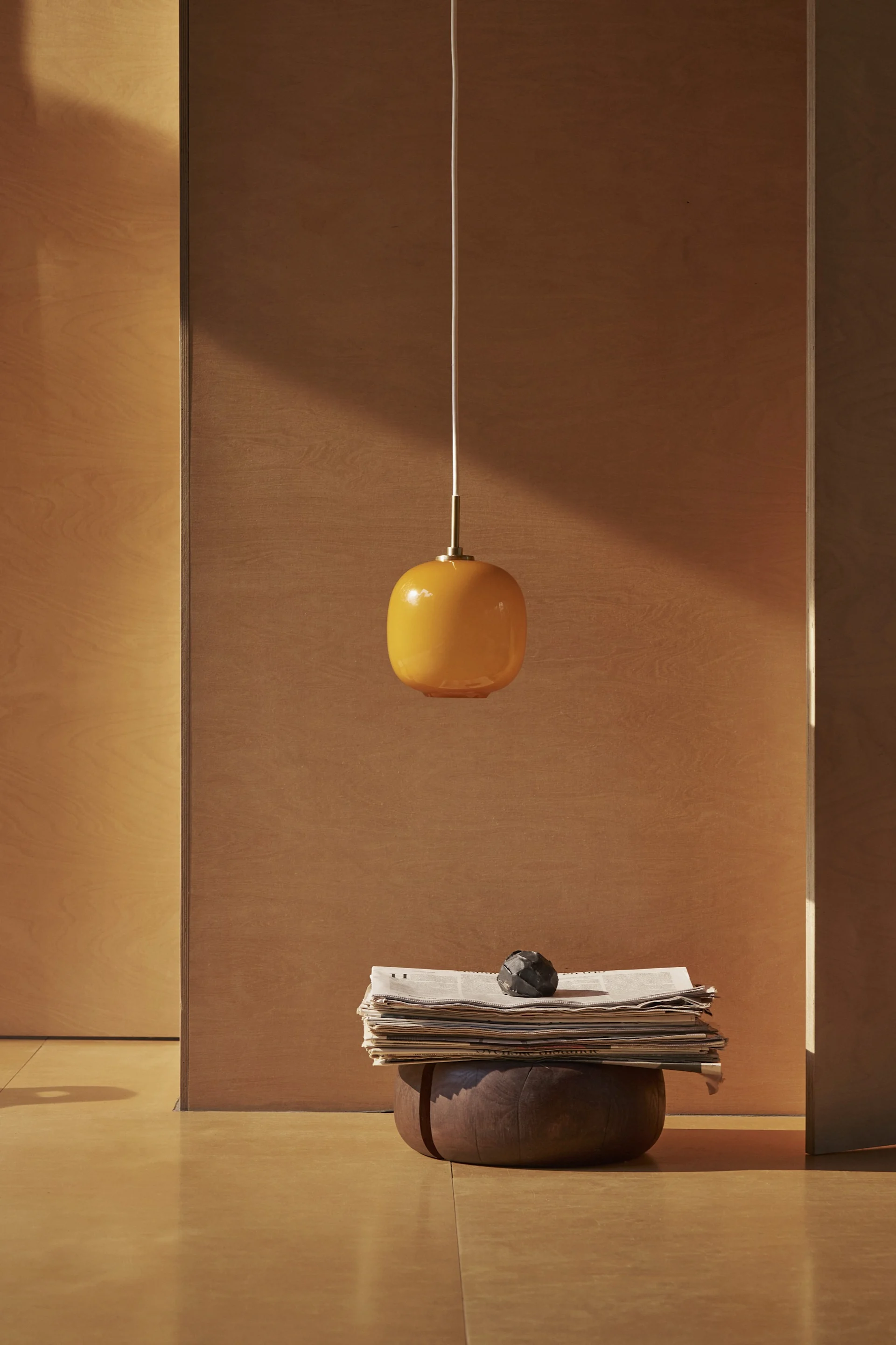 Une suspension ovale orange est suspendue au-dessus d'une pile de journaux et d'un objet décoratif sombre sur une base en bois dans une pièce minimaliste.