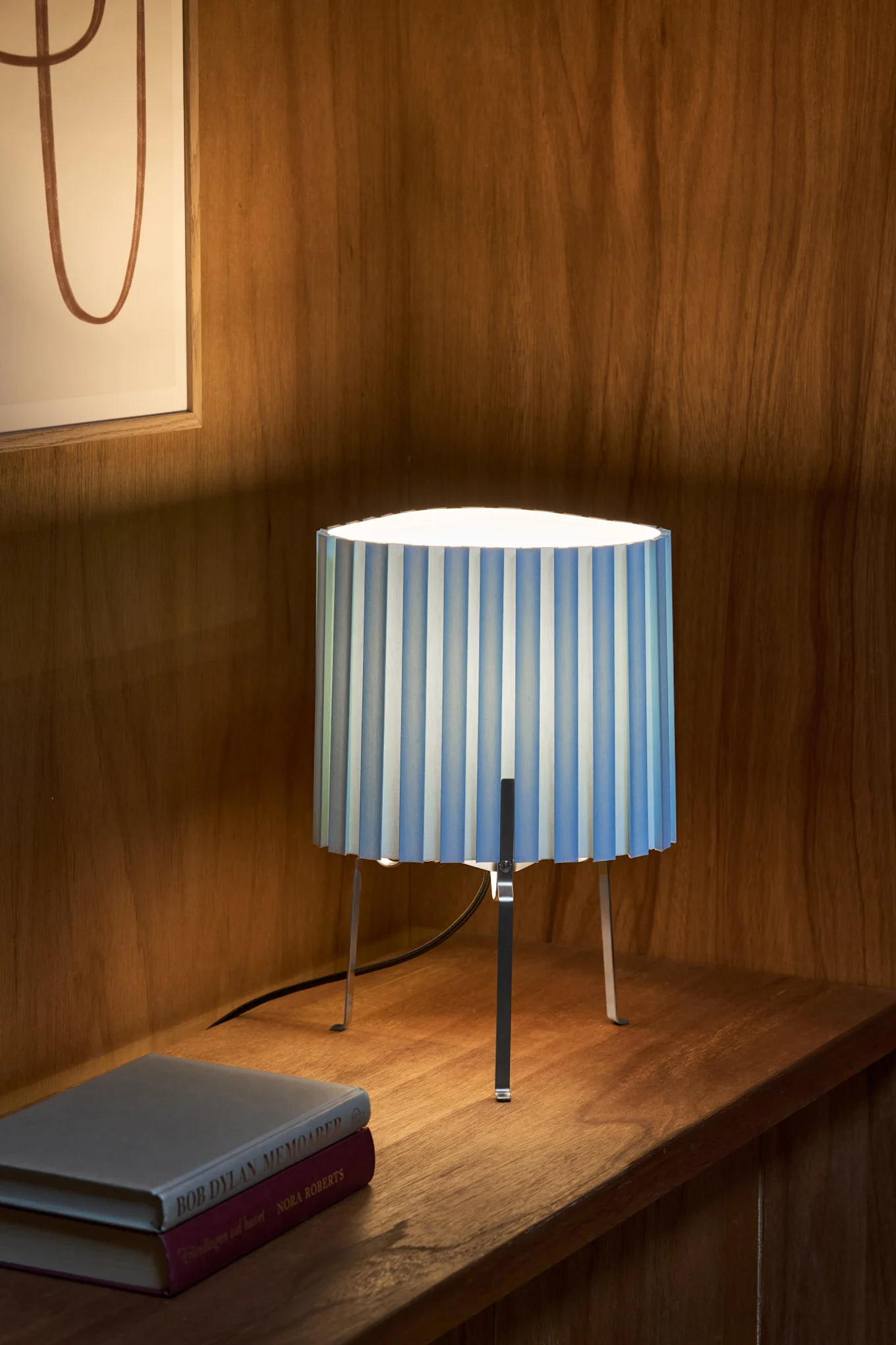 Une lampe de table avec un abat-jour plissé à rayures bleues et blanches et des pieds en métal, posée sur une étagère en bois à côté de deux livres.