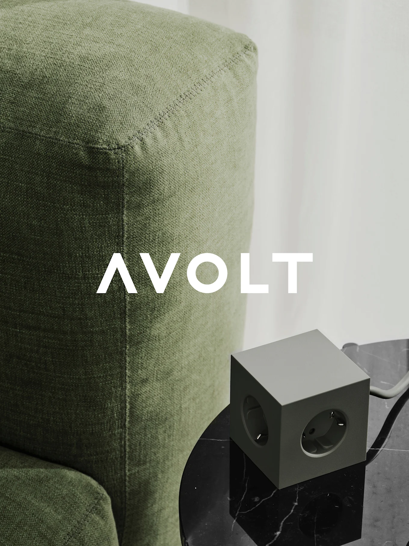Une multiprise cube verte avec trois prises sur une table en marbre noir à côté d'un canapé vert. Logo AVOLT.