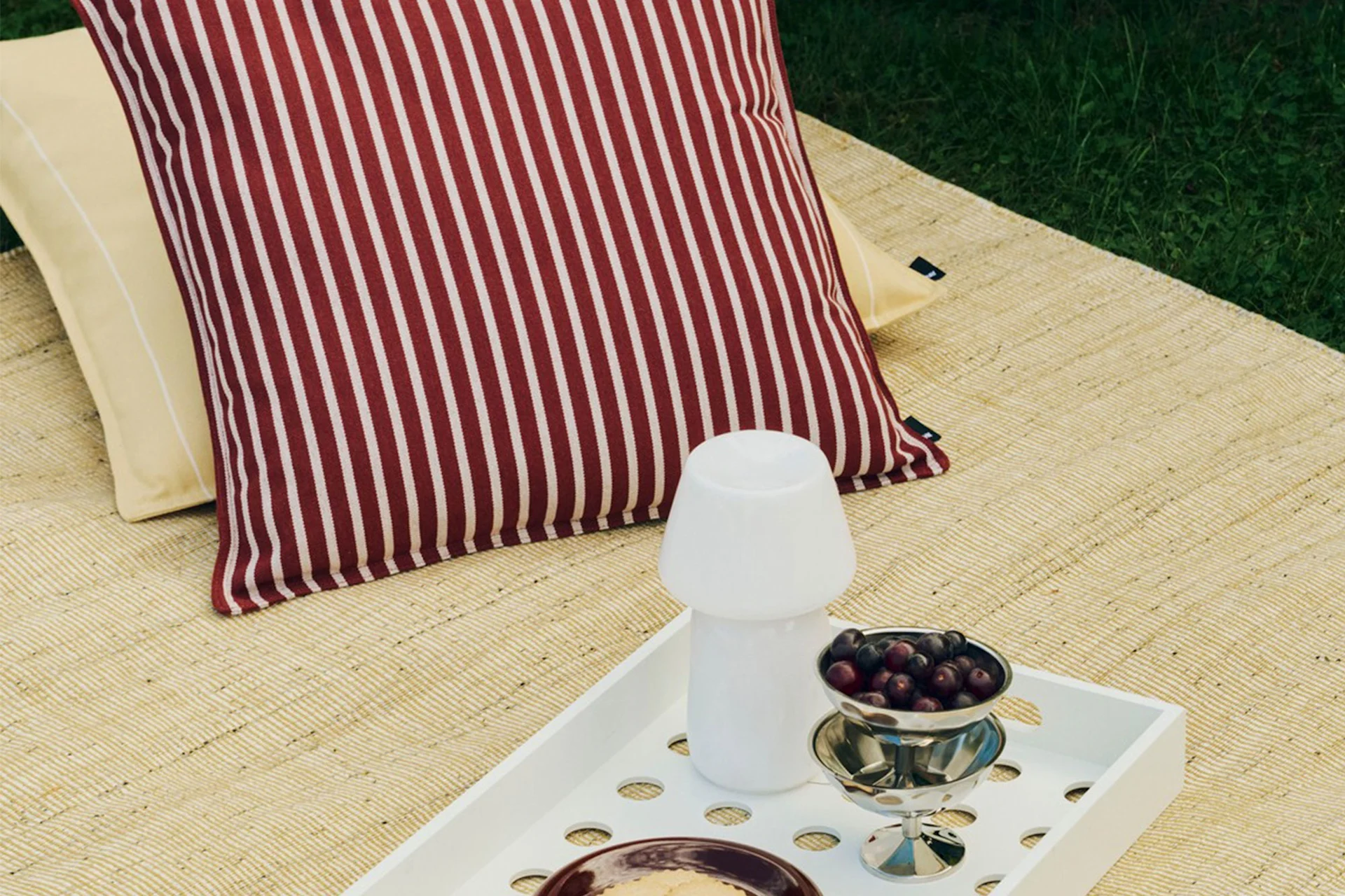 Installation de pique-nique en extérieur avec coussin rayé rouge et blanc, coussin jaune, plateau blanc avec lampe champignon et un bol de baies.