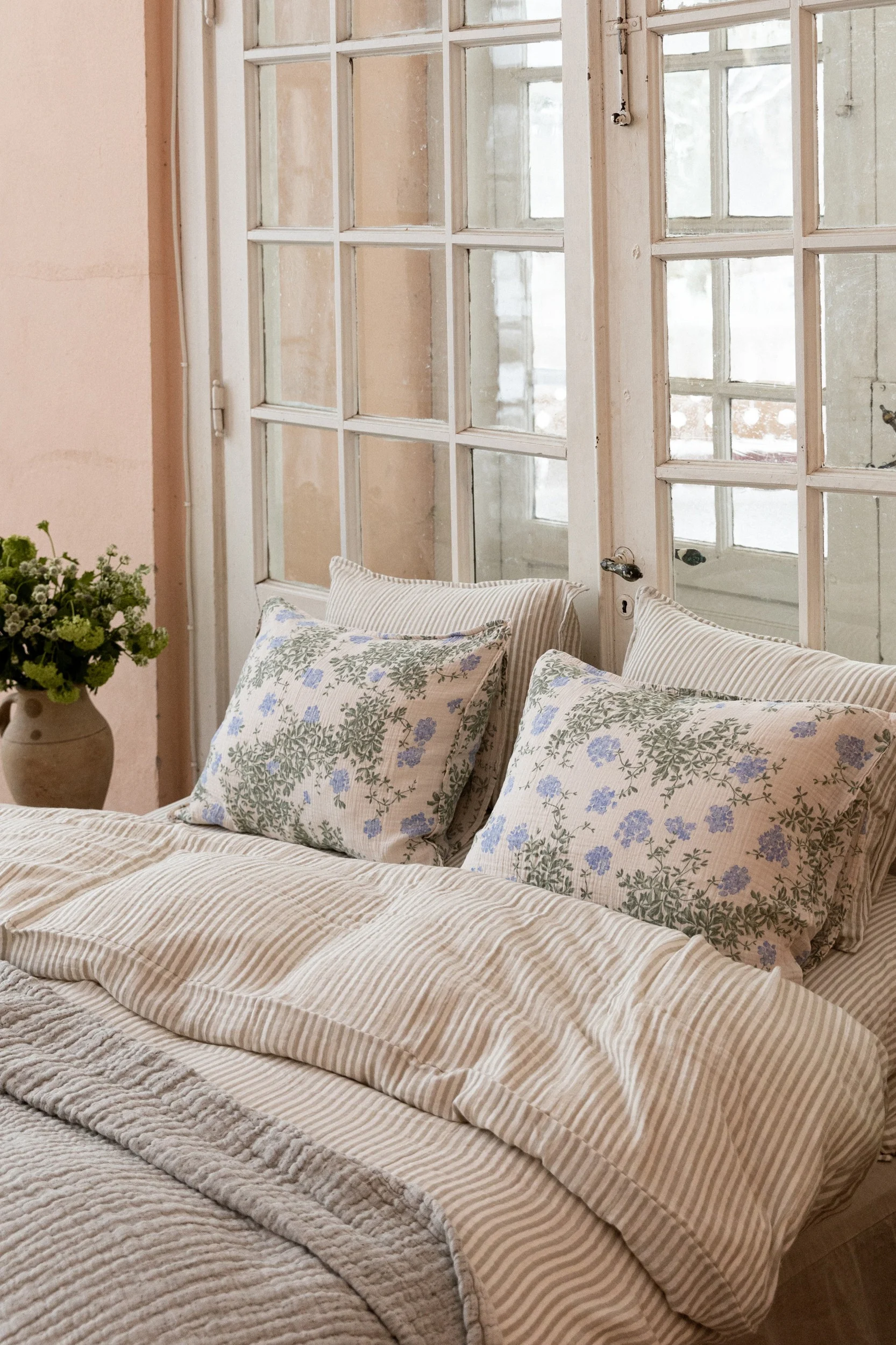 Un lit avec une couette rayée et deux oreillers à motifs floraux bleus, devant une fenêtre à carreaux. Un vase de verdure est à gauche.
