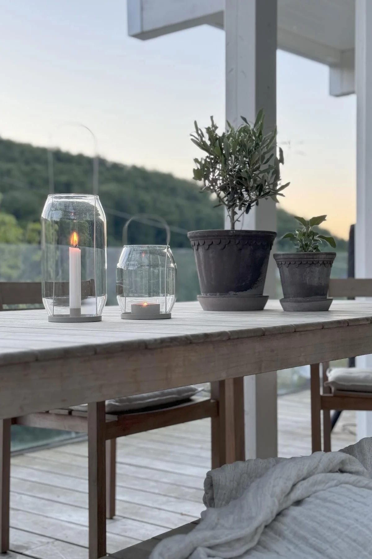 Deux lanternes en verre allumées et deux plantes en pot sur une table en bois extérieure au crépuscule.