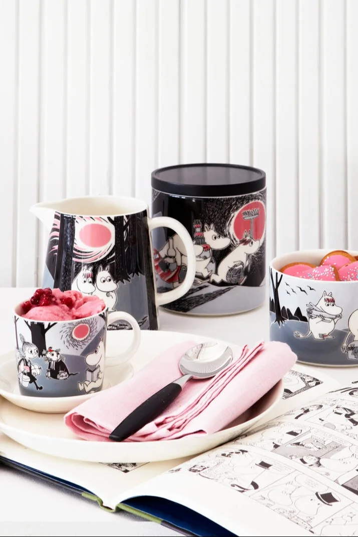 La collection Moomin Adventure, composée d'une tasse, d'une assiette, d'une cruche, d'une boîte de conserve et d'un bol, est posée sur une table.