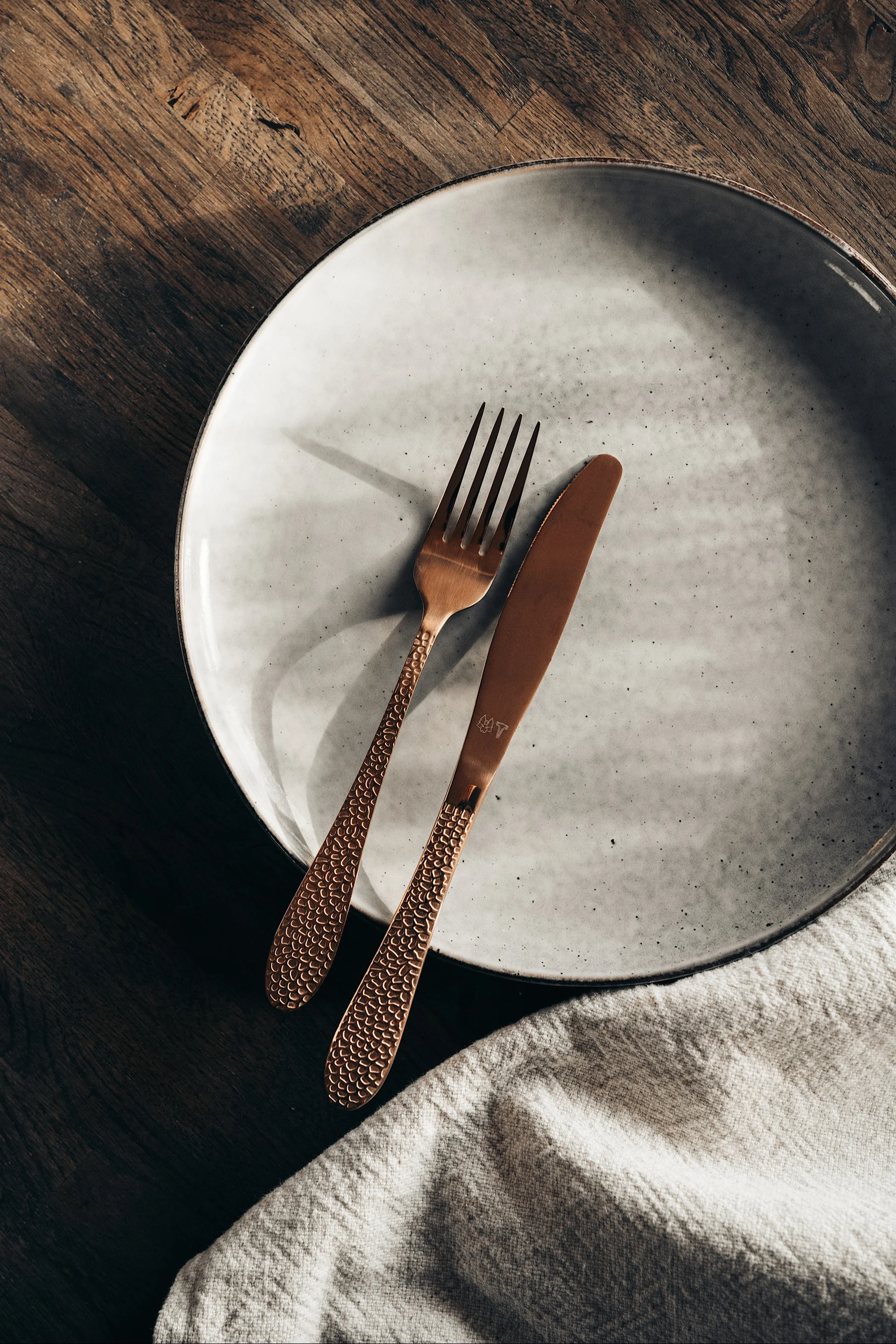 Fourchette et couteau couleur cuivre avec manches texturés sur une assiette en céramique gris clair, à côté d'une serviette en lin sur bois.