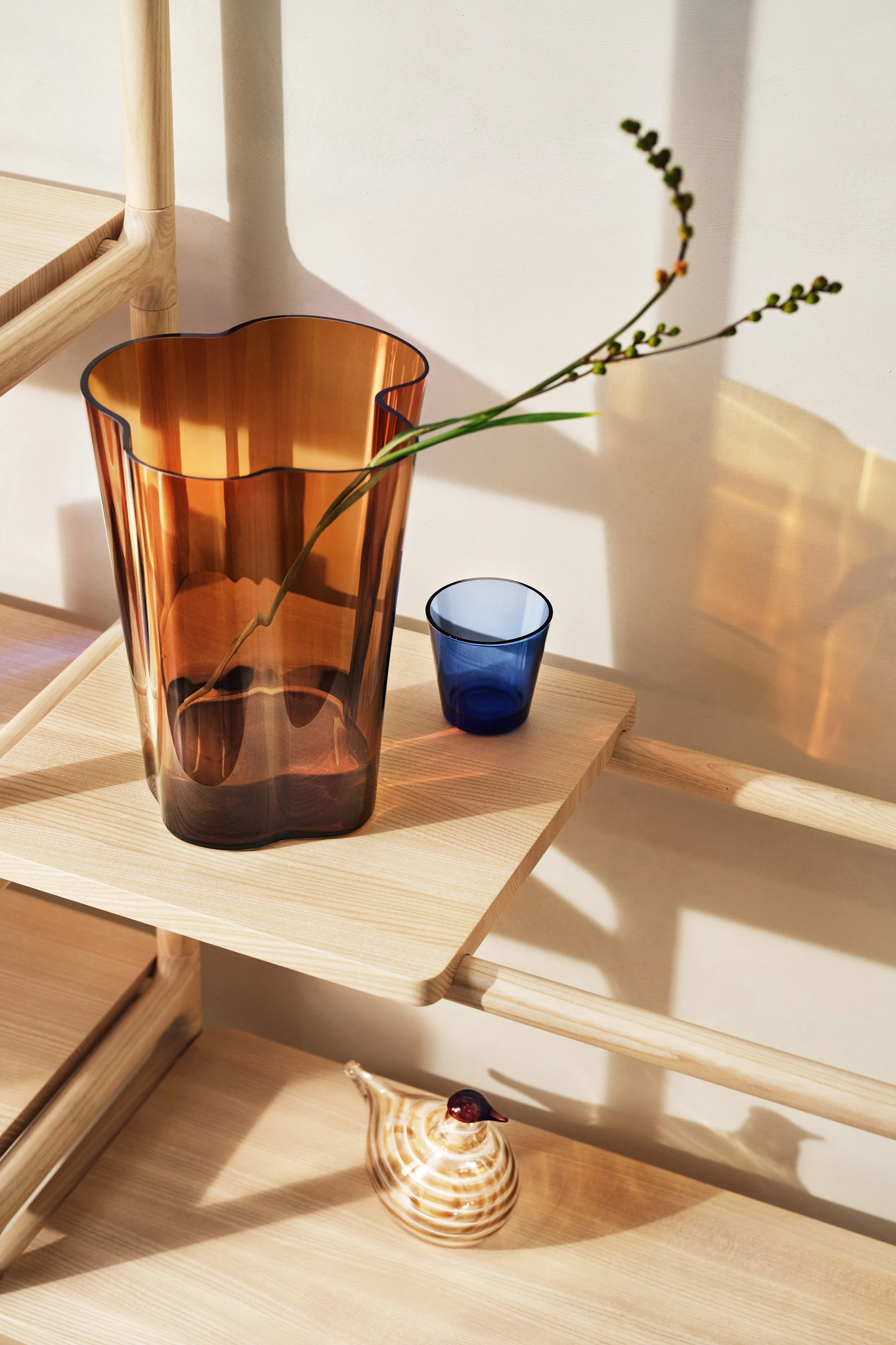 Vase ambré à bord ondulé avec branche, gobelet bleu et oiseau en verre rayé sur étagère en bois clair.