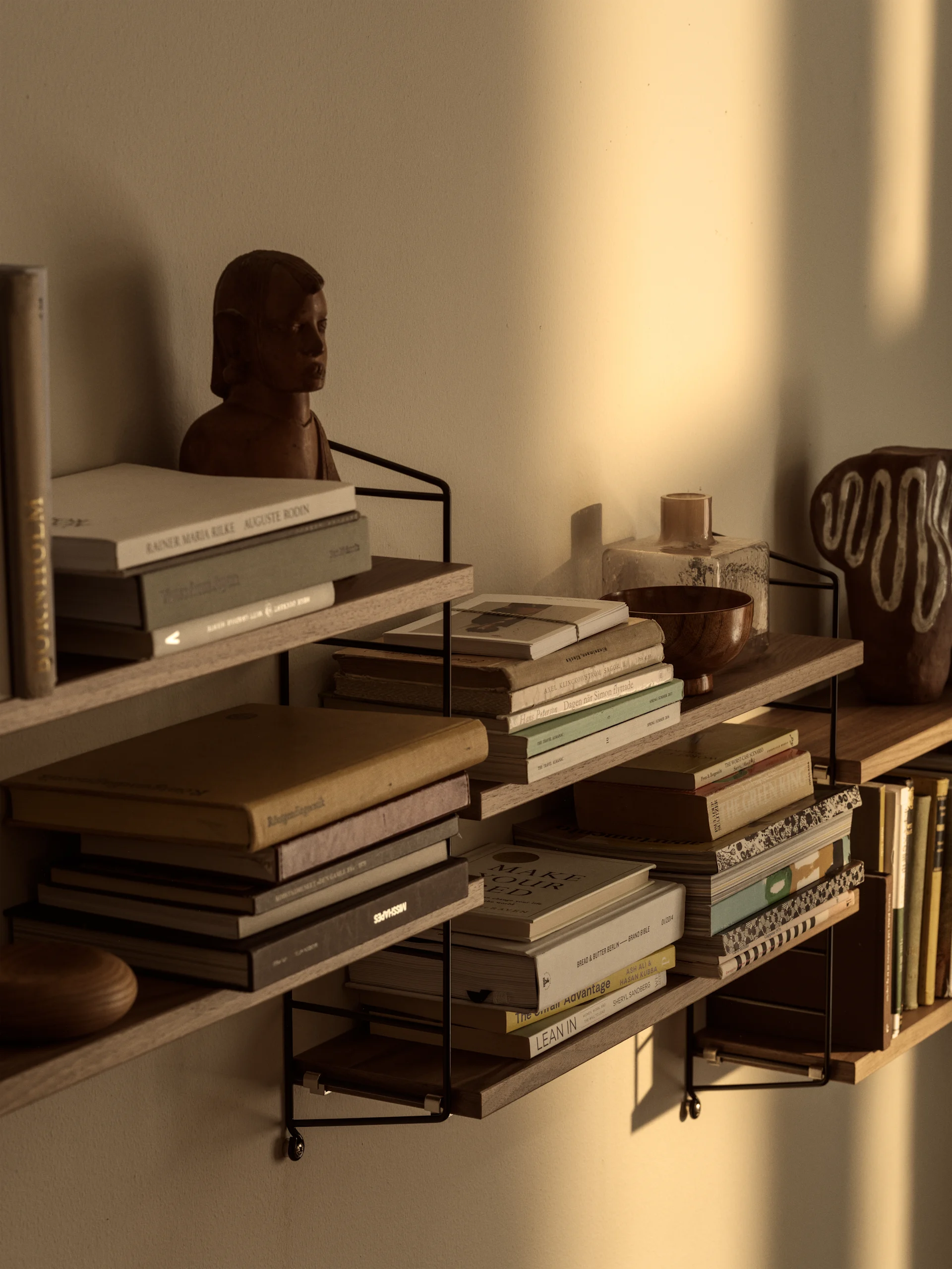 Étagères murales remplies de livres, d'un buste en bois et d'objets décoratifs, éclairées par la chaude lumière du soleil sur le mur.