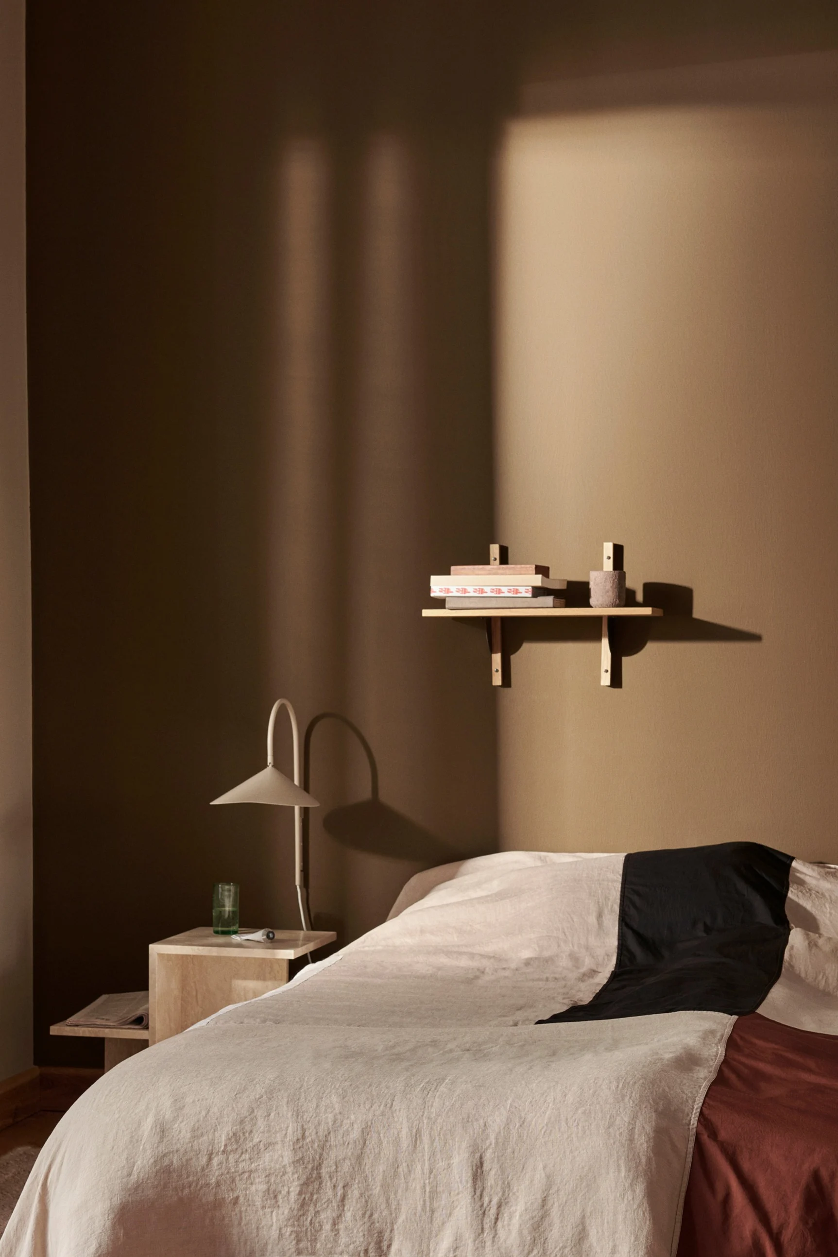 Une chambre avec un lit aux draps beiges, noirs et rouille, une table de chevet avec une lampe et une étagère avec des livres sur un mur marron chaud.