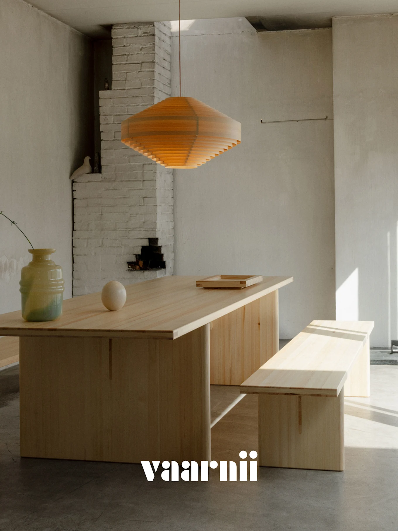 Une salle à manger minimaliste avec une longue table en bois clair, un banc assorti, une suspension en bois superposé, un vase et une sphère. Logo Vaarnii.