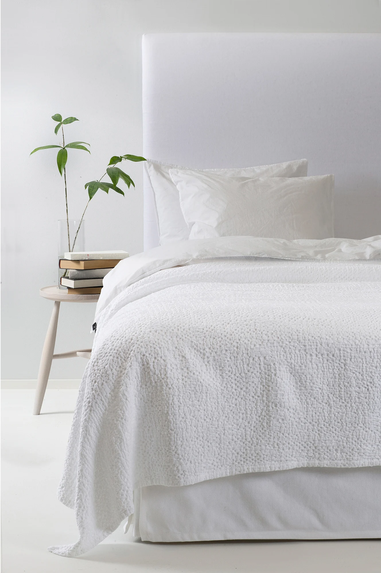 Lit blanc avec couette texturée, oreillers, tête de lit rembourrée, table d'appoint en bois avec livres et plante verte.
