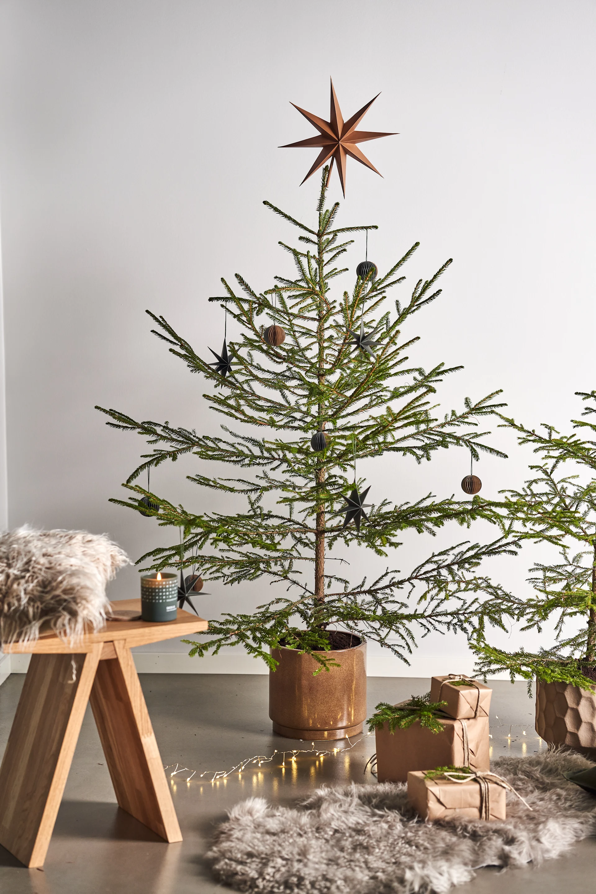 Décorez le sapin de Noël avec des décorations de sapin de Noël pour 2021 dans 4 styles différents selon les tendances Nest - Nurture, Share, Boost et Cultivate. Ici, vous voyez un tabouret Angle de Muubs avec une bougie parfumée Skandinavisk dans la senteur Forest à côté de paquets sur un tapis en peau de mouton. 