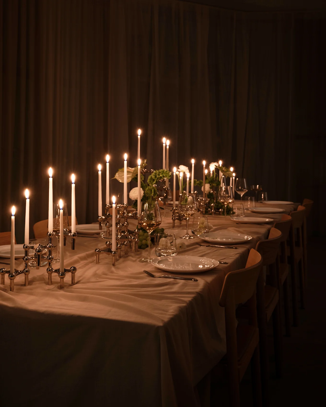 Longue table à manger avec bougies allumées dans bougeoirs argentés, assiettes blanches et verres. Lueur chaude dans pièce sombre.
