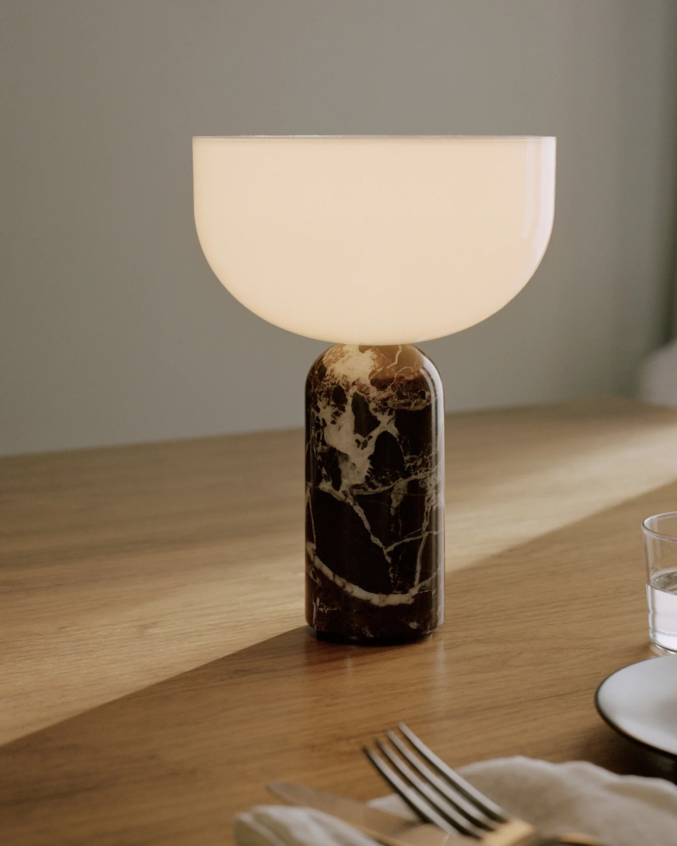 Lampe de table avec une base cylindrique en marbre foncé veiné de blanc et un abat-jour semi-circulaire en verre blanc lumineux, posée sur une table en bois avec des couverts et un verre.