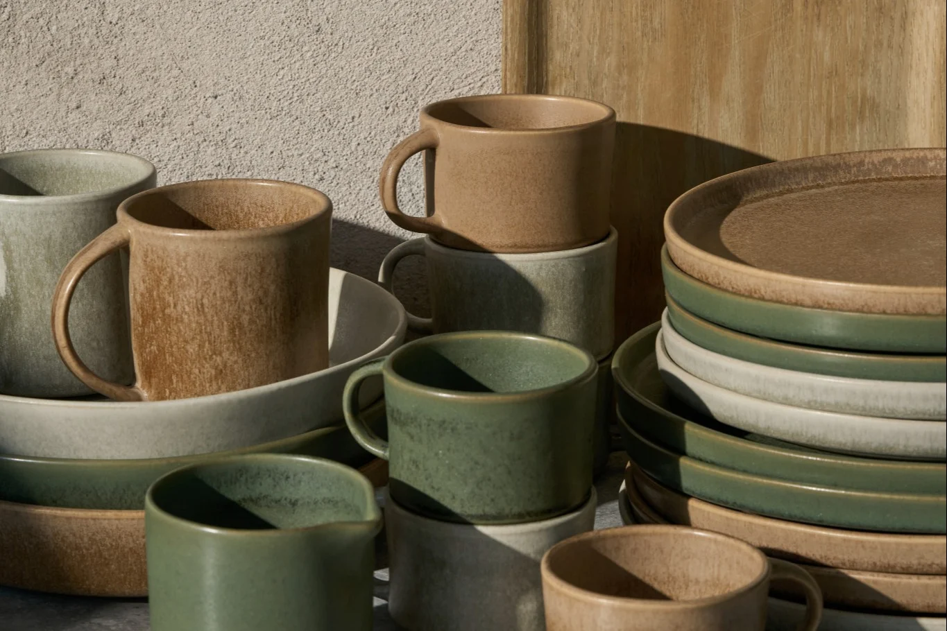Tasses, bols et assiettes en céramique empilés en beige, gris clair et vert sauge, avec une finition rustique.