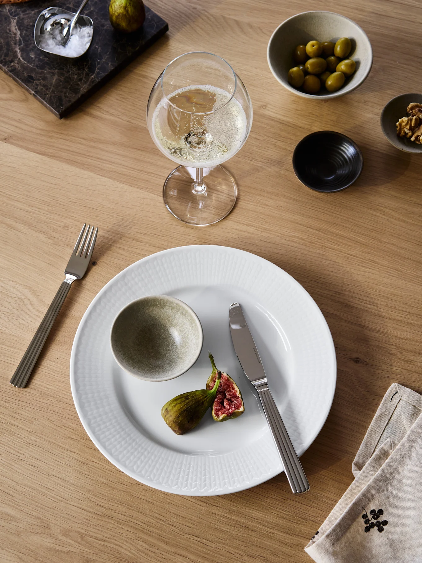 Table à manger avec assiette blanche, figue coupée, fourchette, couteau, vin pétillant, olives, noix, craquelins et sel sur une planche en marbre.