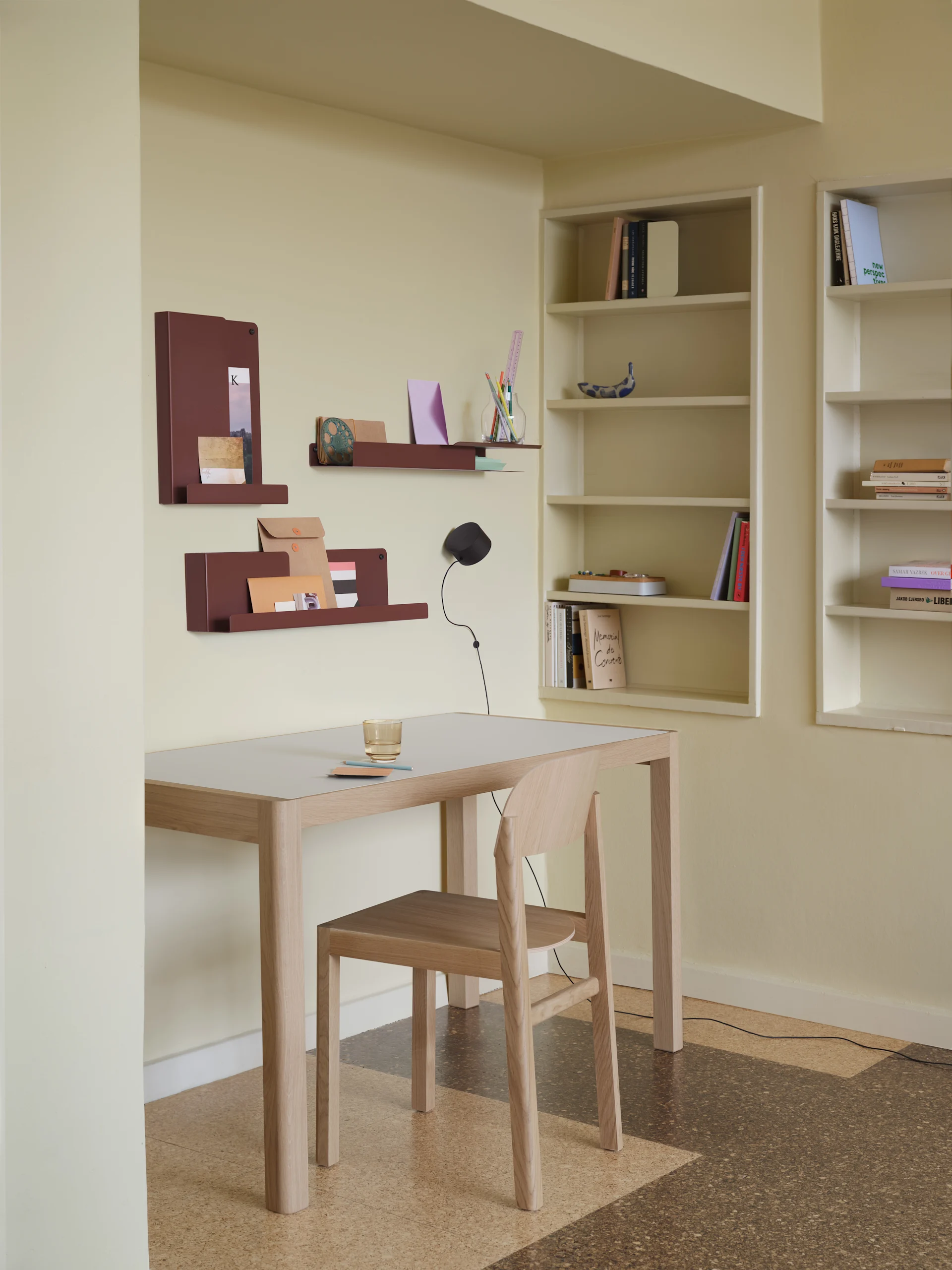 Un aménagement de bureau minimaliste avec un bureau en bois clair, une chaise, des organisateurs muraux marron rougeâtre et des bibliothèques encastrées.