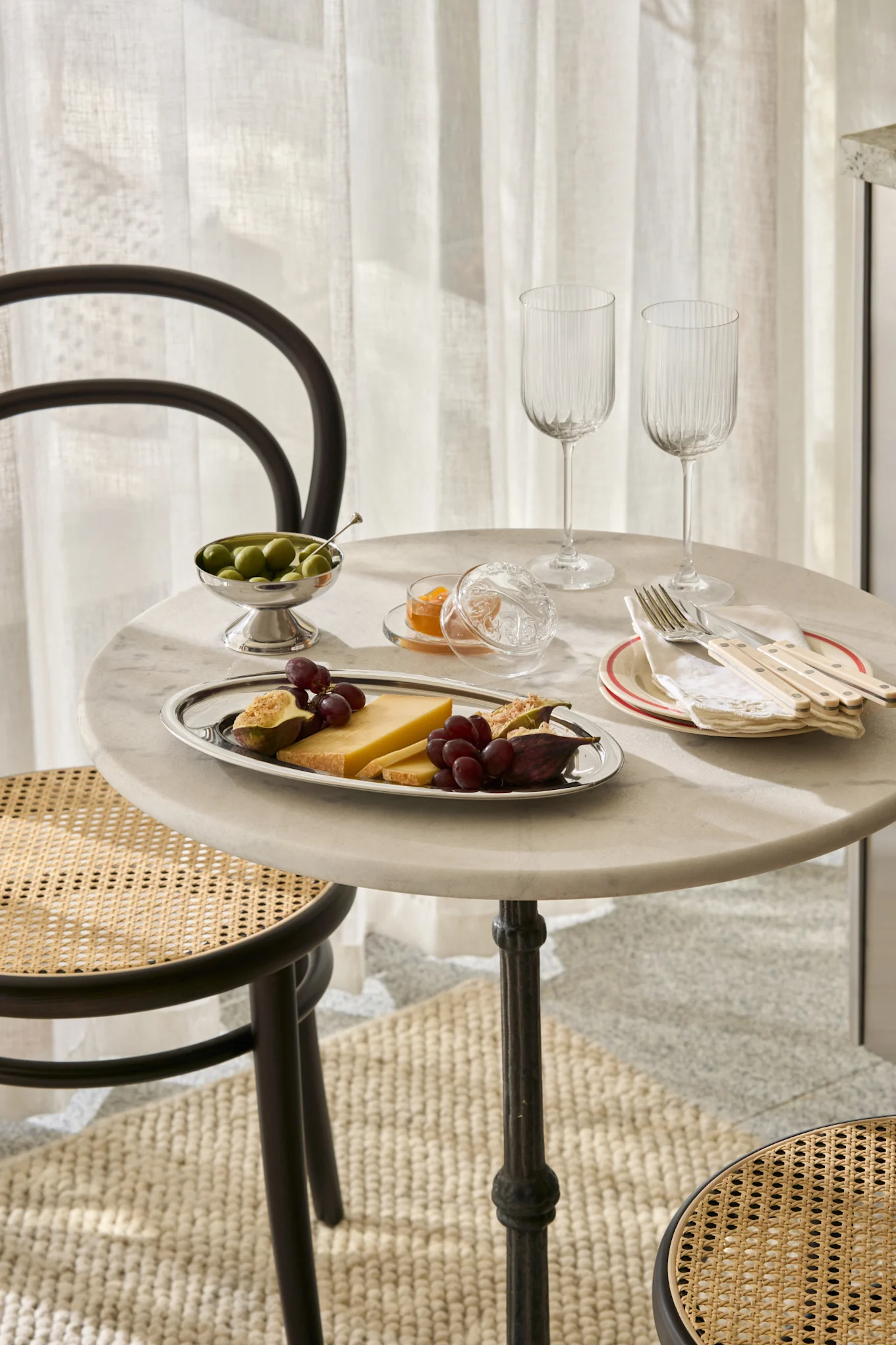 Une table en marbre dressée avec fromage, figues, raisins, olives, confiture, verres et couverts, avec des chaises en rotin.