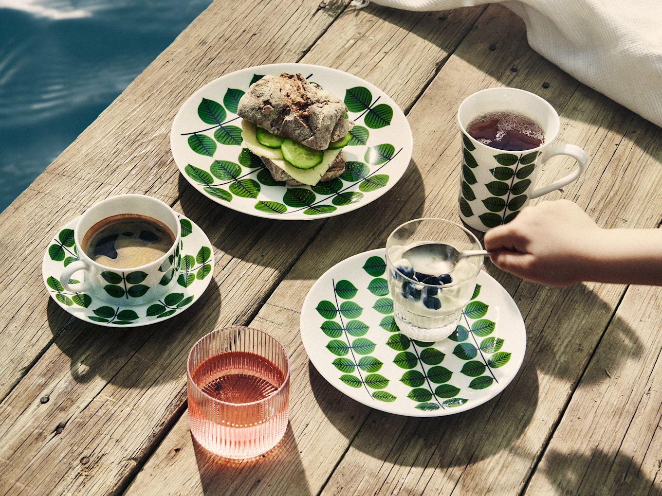 Petit-déjeuner sur jetée en bois: vaisselle à feuilles, sandwich, café, thé, yaourt aux myrtilles et boisson rose.