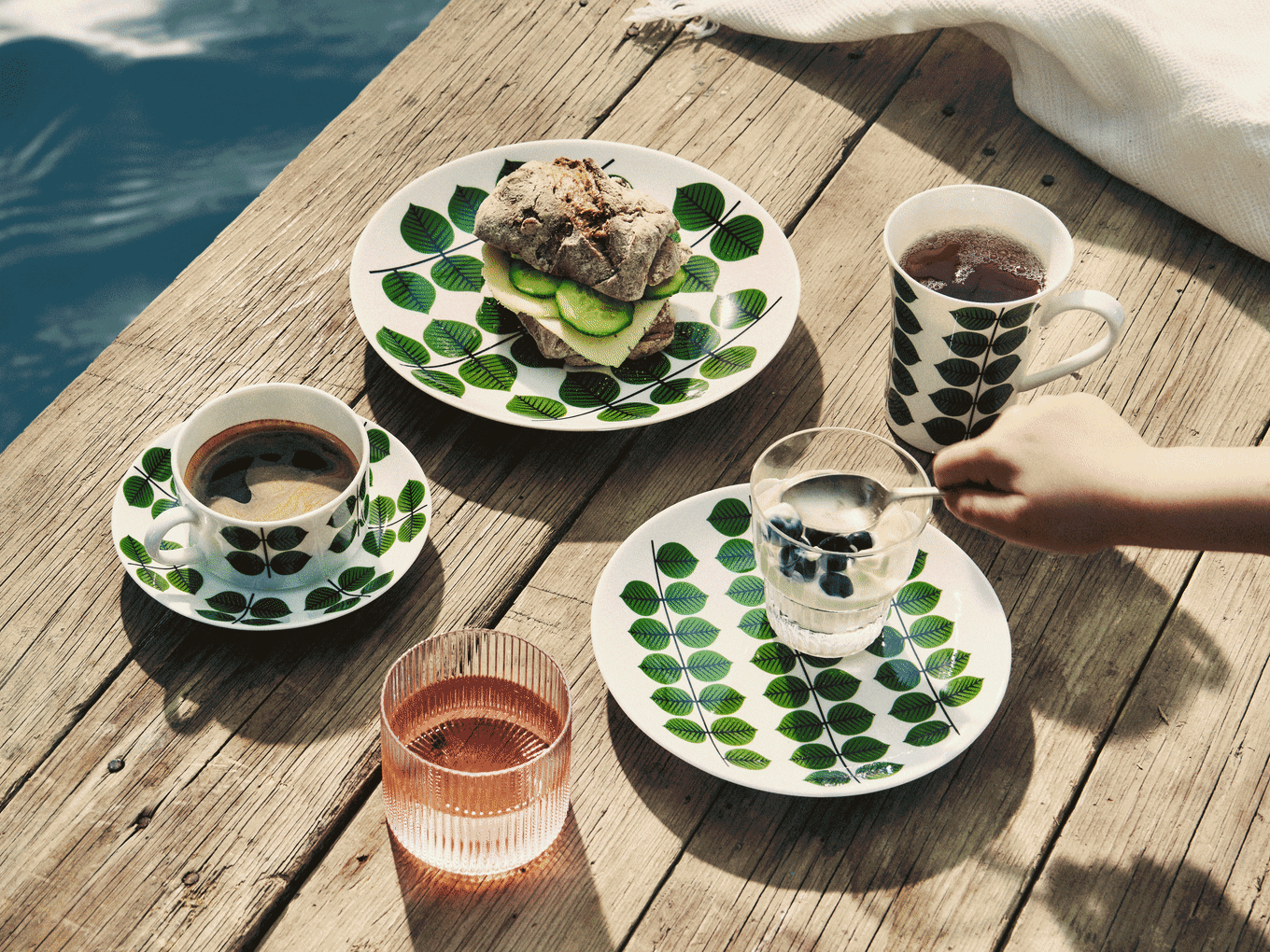 Petit-déjeuner sur jetée en bois: vaisselle à feuilles, sandwich, café, thé, yaourt aux myrtilles et boisson rose.