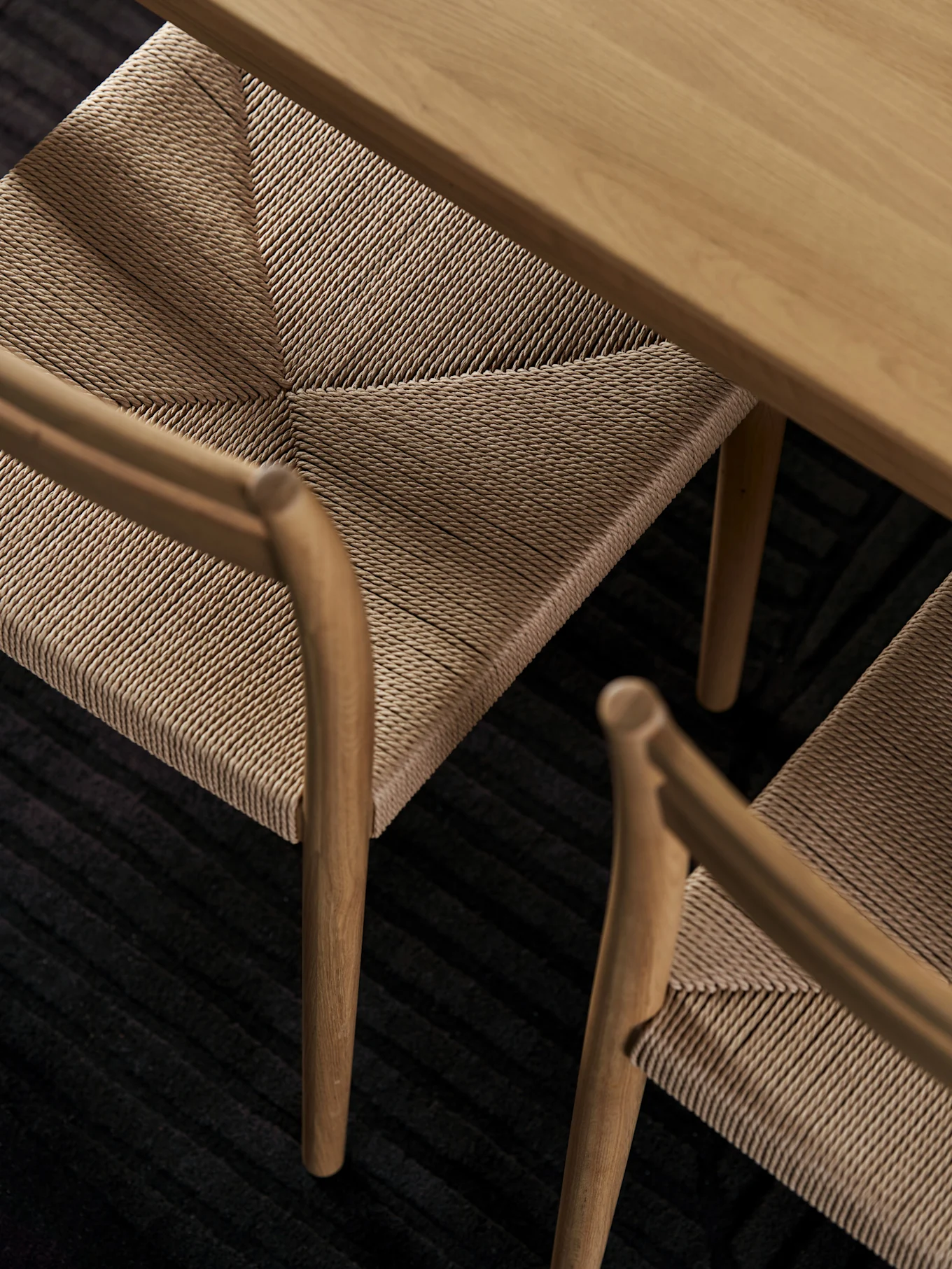 Vue plongeante d'une table en bois clair et de deux chaises aux assises tressées sur un tapis nervuré foncé.