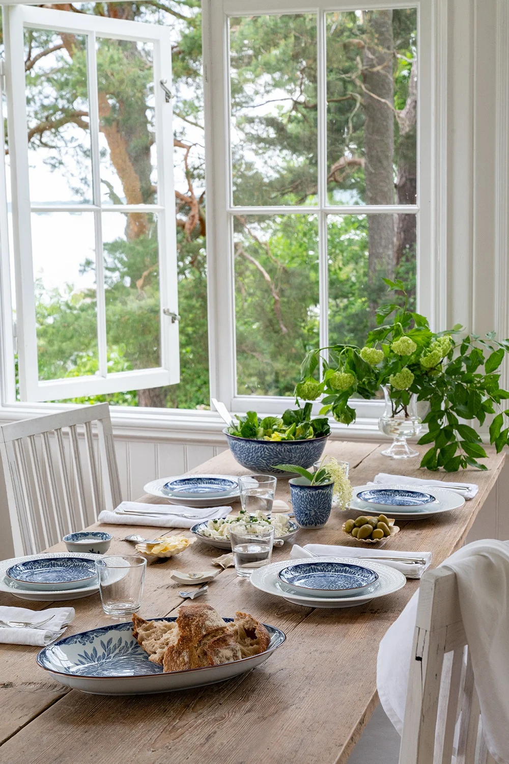 Une table à manger rustique en bois dressée avec des assiettes à motifs bleus et blancs, des verres d'eau, du pain, de la salade et des olives, à côté d'une fenêtre ouverte donnant sur des arbres verts et de l'eau.