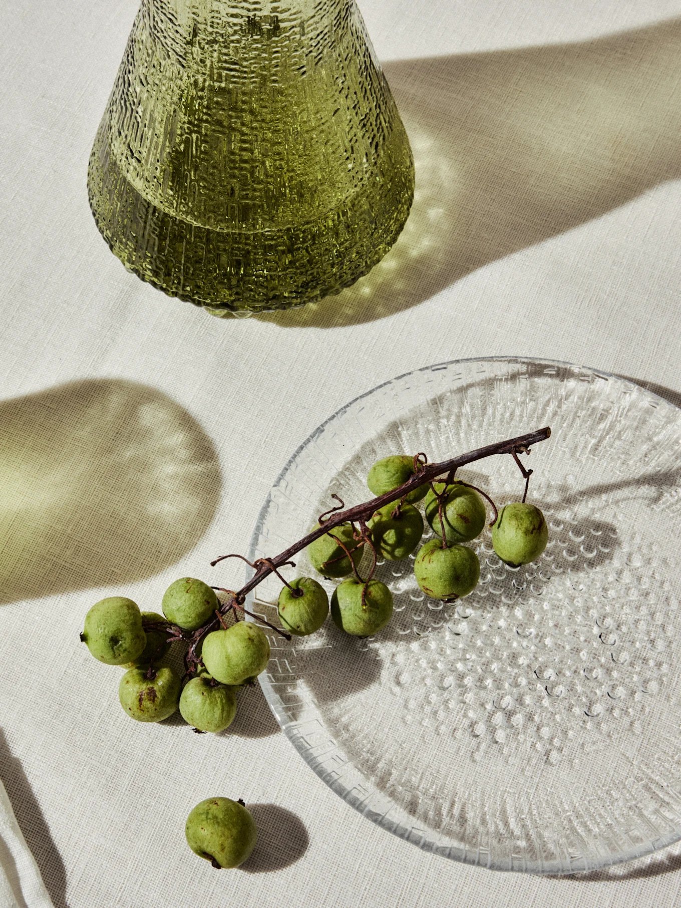 Vase en verre vert texturé et une assiette transparente avec de petits fruits verts sur une branche.