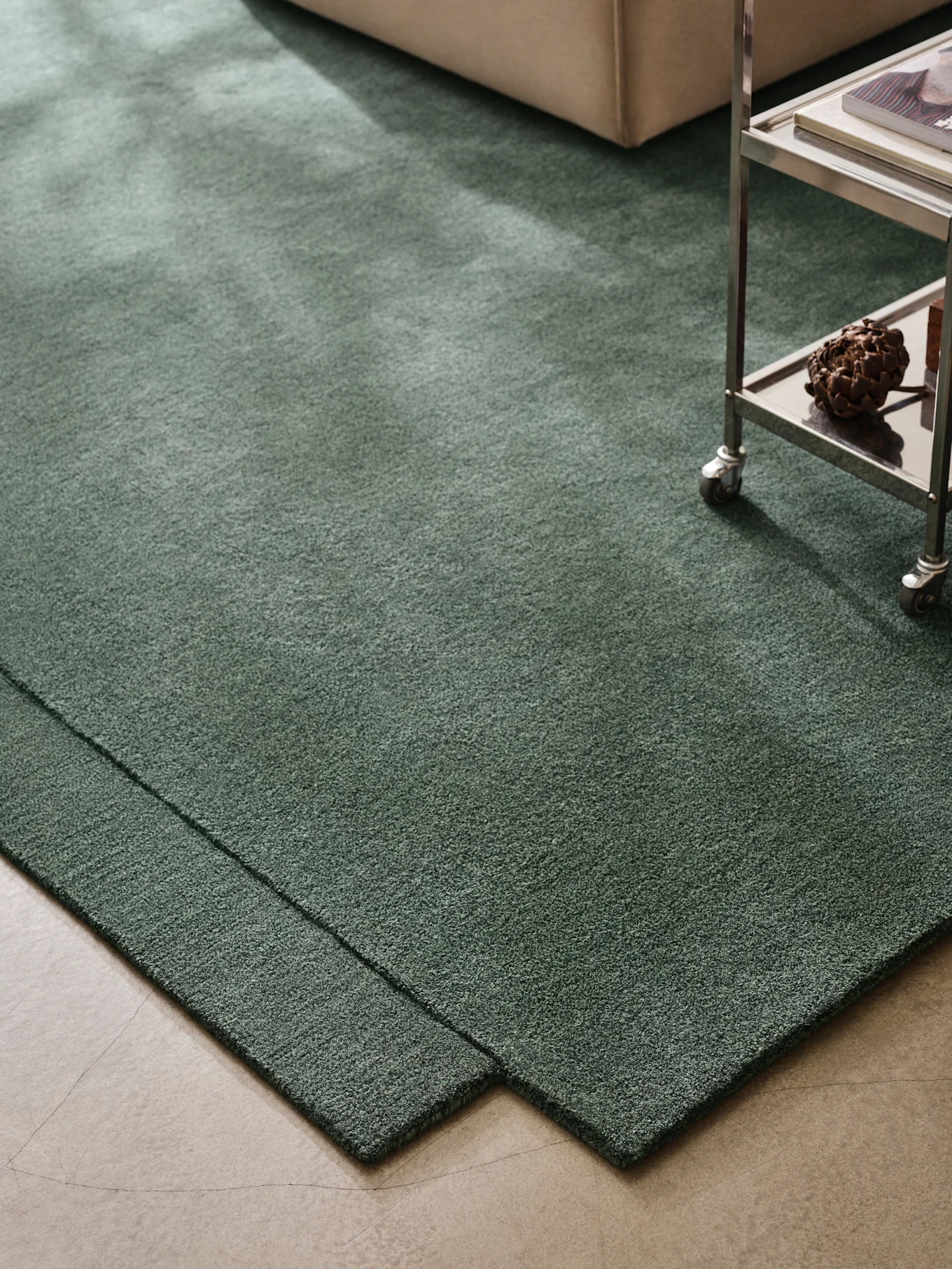Tapis vert foncé à poils longs sur sol en béton, avec un canapé beige et un chariot métallique.