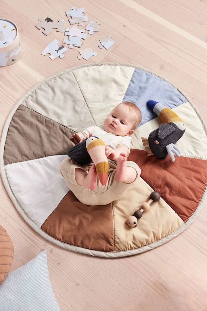 La peluche Trine Tourca de OYOY est allongée à côté d'un bébé sur le tapis pour enfants Moni. La peluche est fabriquée exclusivement à partir de matériaux recyclés, ce qui en fait non seulement un excellent cadeau pour une baby shower, mais aussi un cadeau durable.