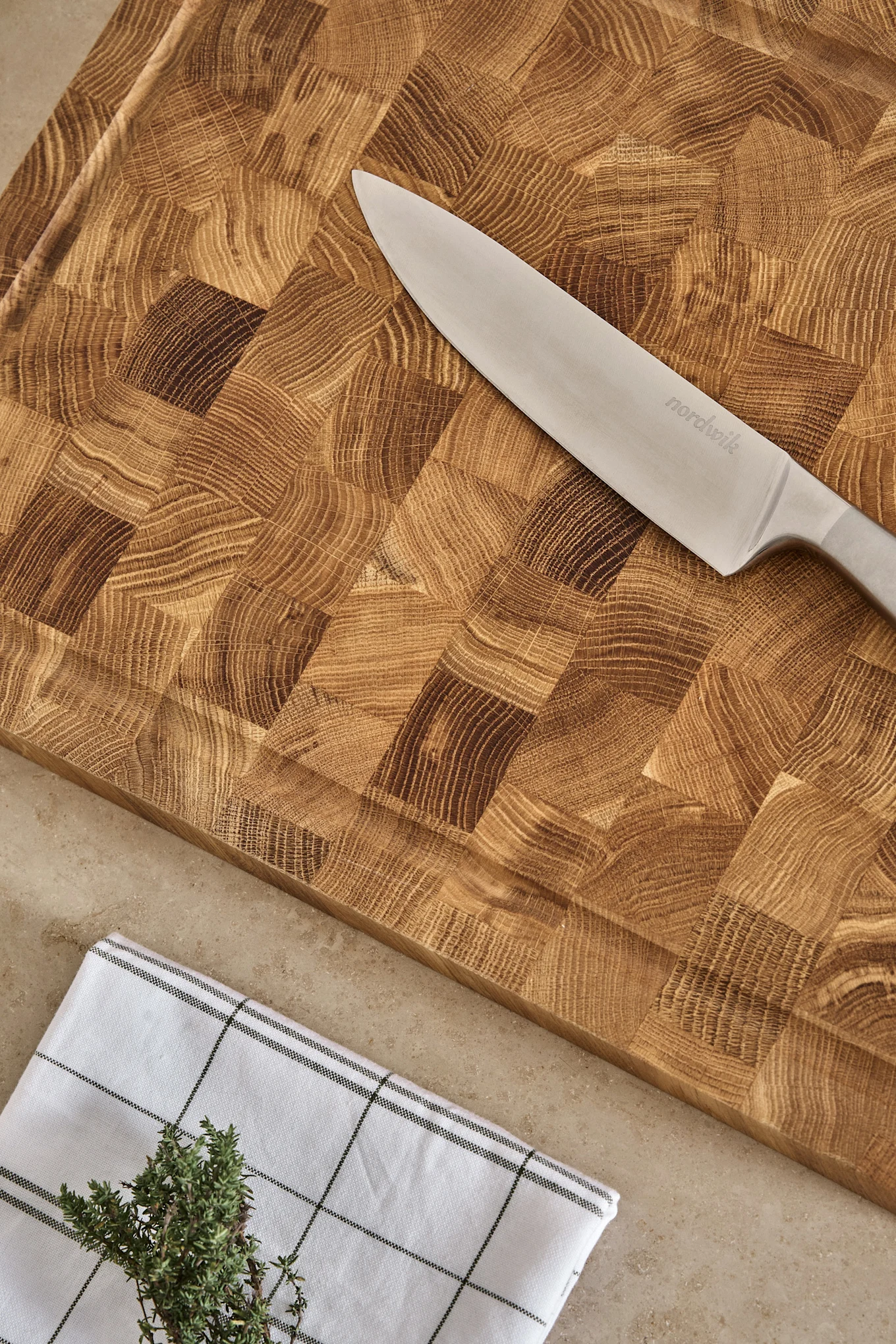 Un couteau de cuisine Nordwik posé sur une planche à découper en bois massif, avec un torchon à carreaux et du thym frais.