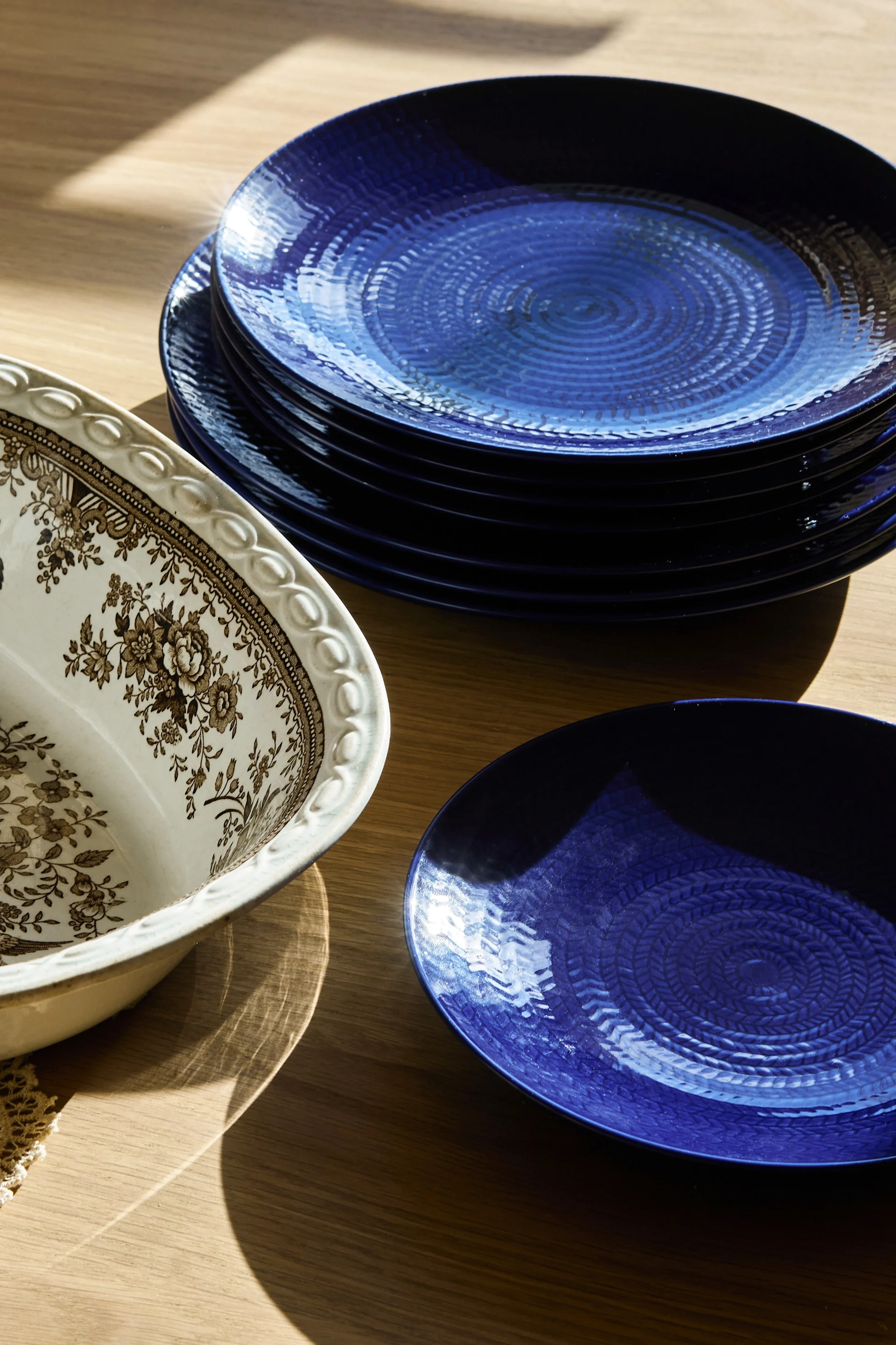 Une pile d'assiettes bleu foncé texturées, accompagnées d'un saladier orné d'un motif floral blanc, marron et crème, posée sur une table en bois.