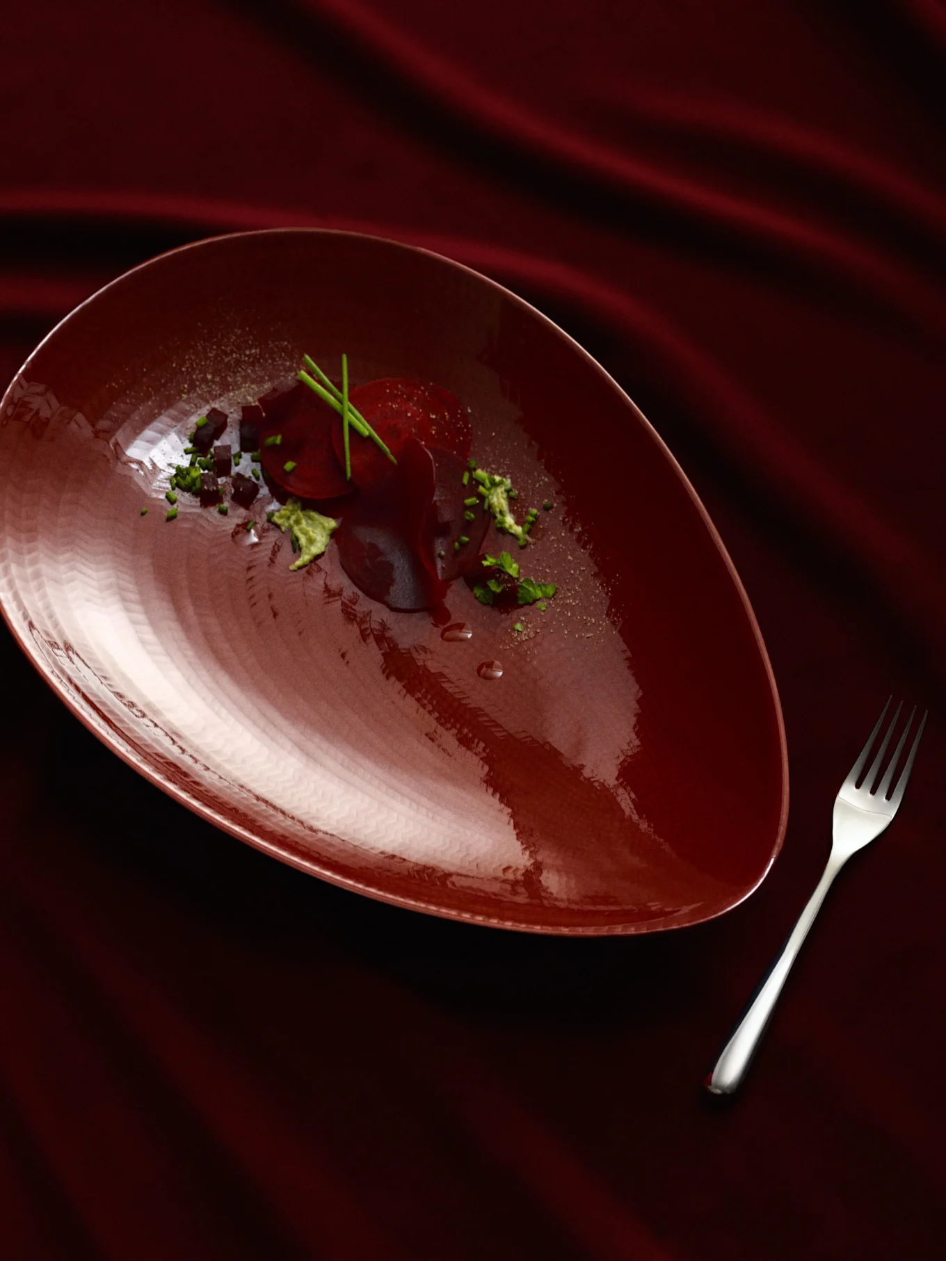 Une assiette rouge foncé en forme de poire avec un plat de betteraves, garnie d'herbes vertes et de cubes foncés, à côté d'une fourchette argentée sur un tissu rouge foncé drapé.