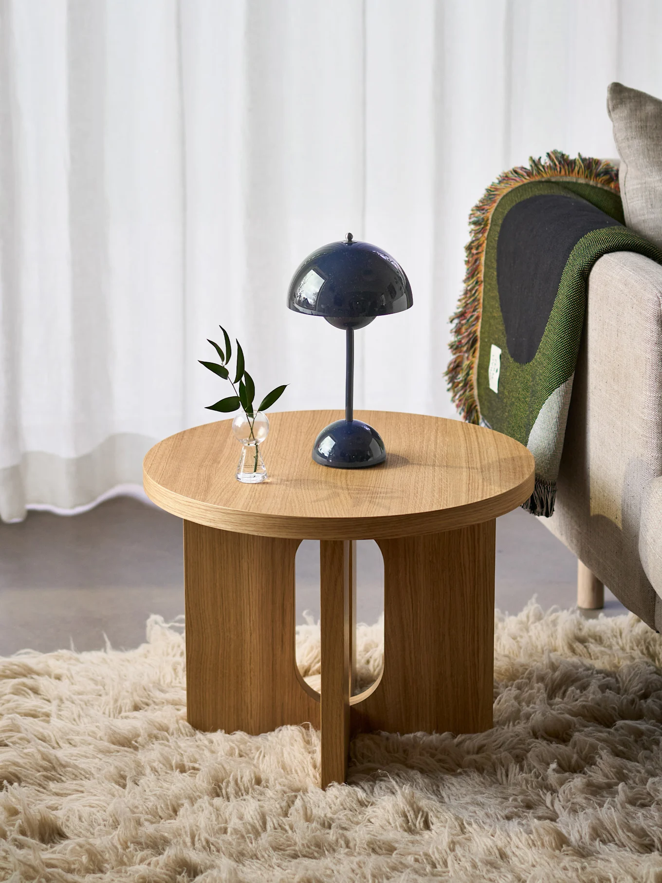 Table ronde en bois avec lampe bleue et plante, à côté d'un fauteuil avec couverture verte, sur un tapis moelleux.