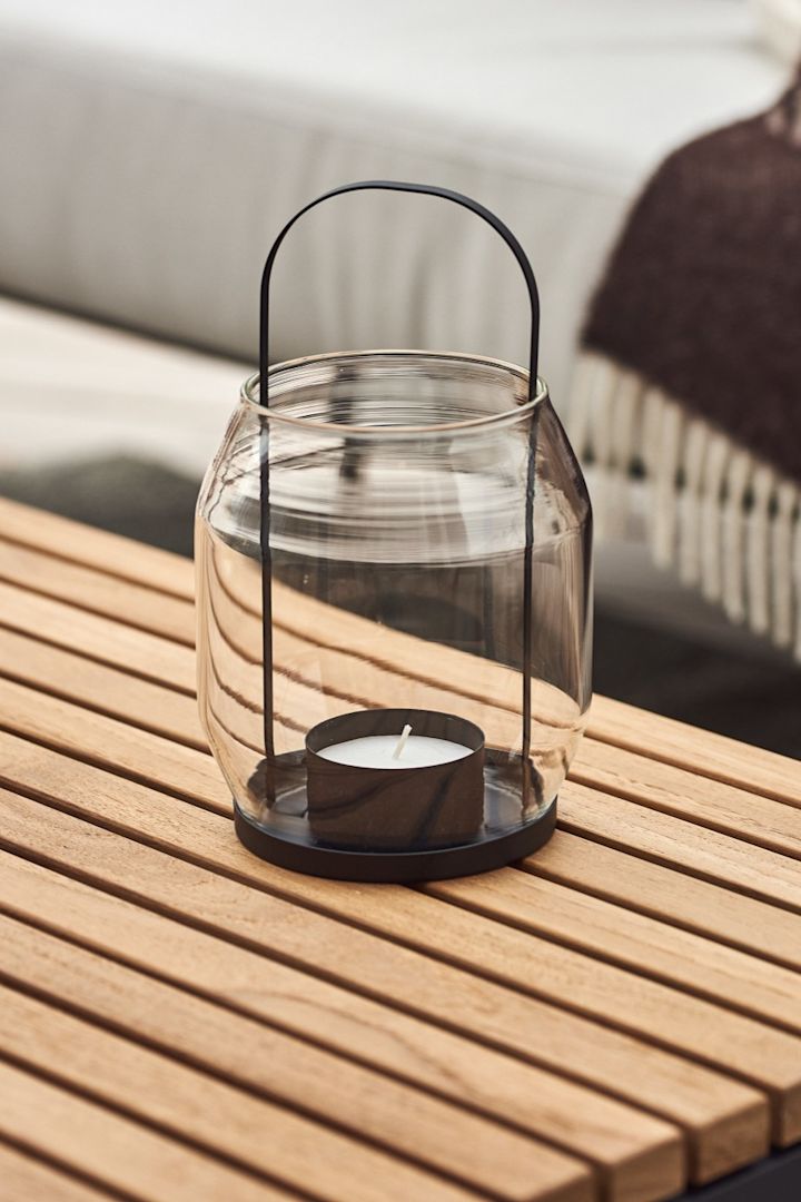 Lanternes et lampes-tempête pour l’extérieur – ici, vous voyez la lanterne ERNST en verre et métal noir, posée sur une table de balcon en bois.