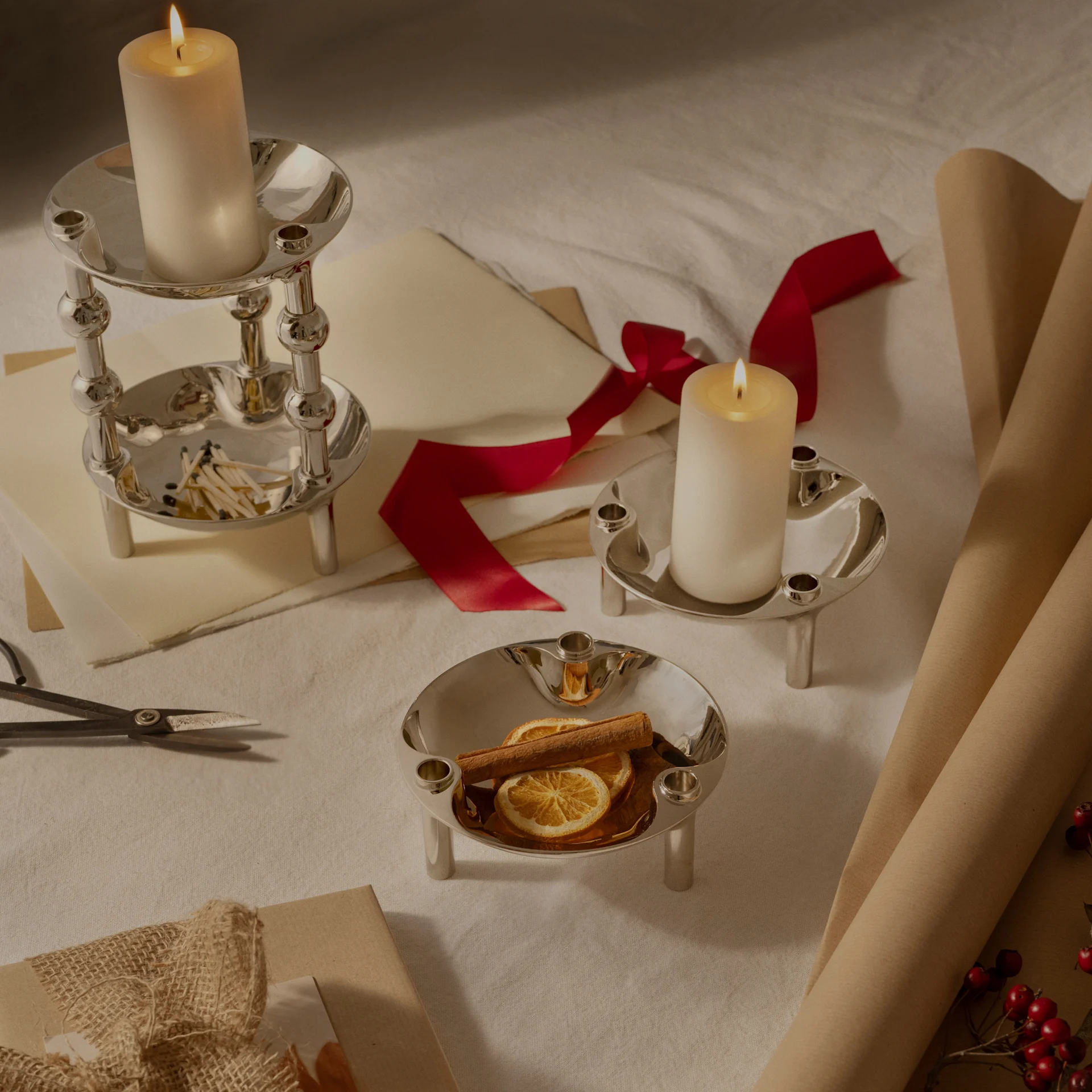 Trois bougeoirs argentés, deux avec des bougies piliers allumées, un avec des oranges séchées et de la cannelle, arrangés avec du papier cadeau et un ruban.