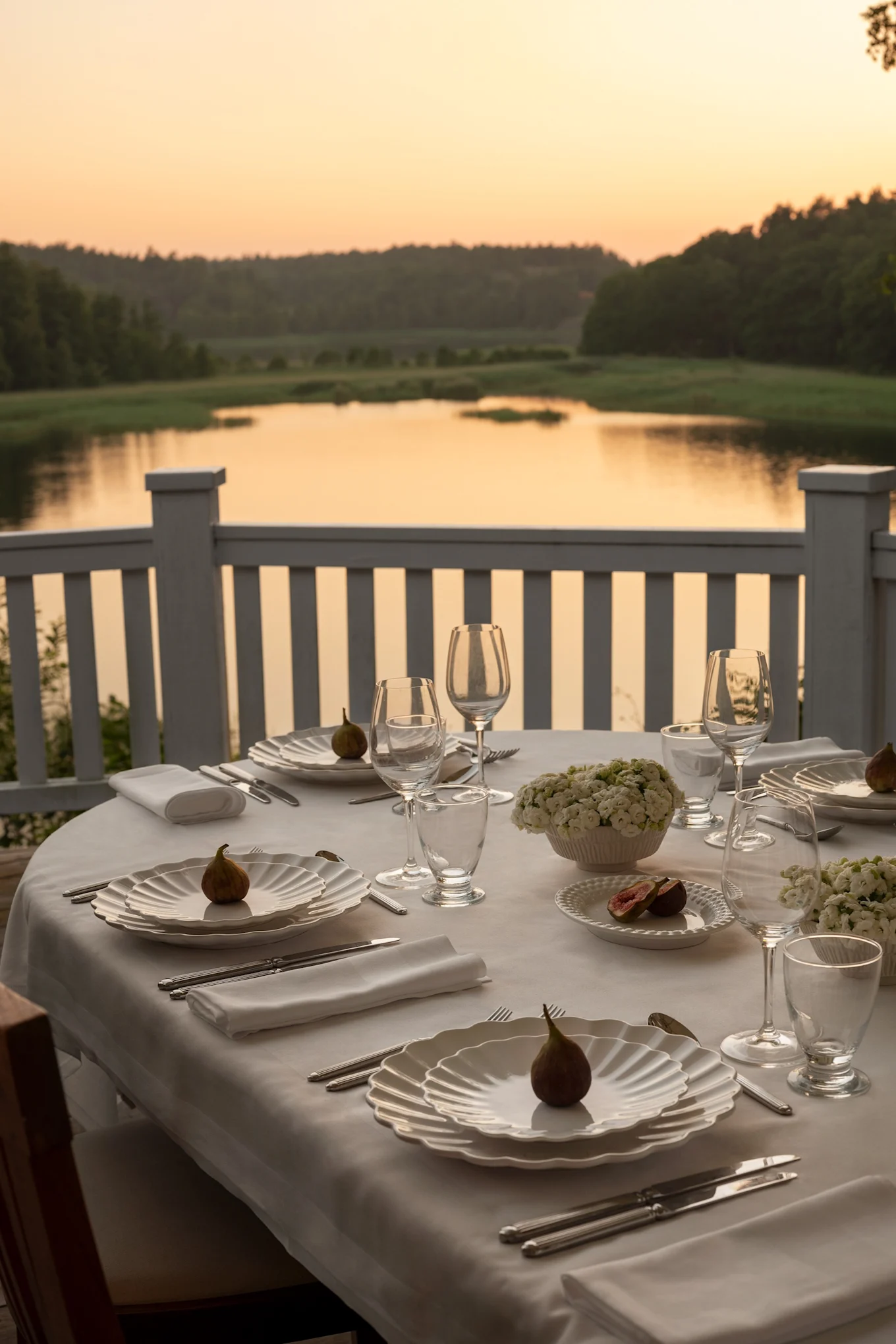 Une table au coucher du soleil sur une terrasse surplombant un lac. La table est dressée en blanc avec des assiettes de la collection Oyster de Mateus.