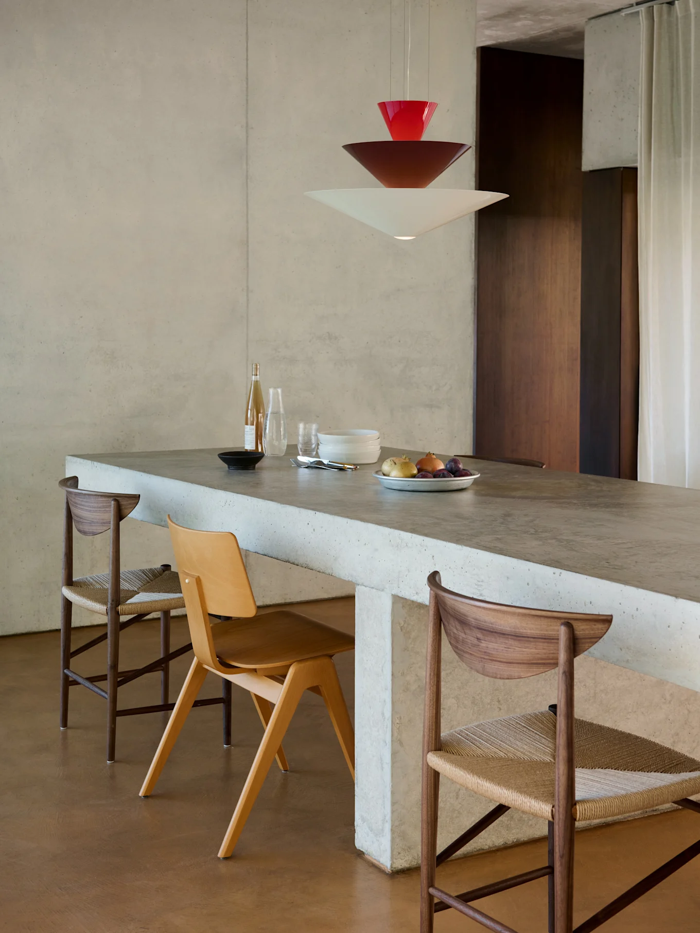 Espace repas moderne avec îlot en béton, trois chaises et suspension empilée rouge, marron et blanche.
