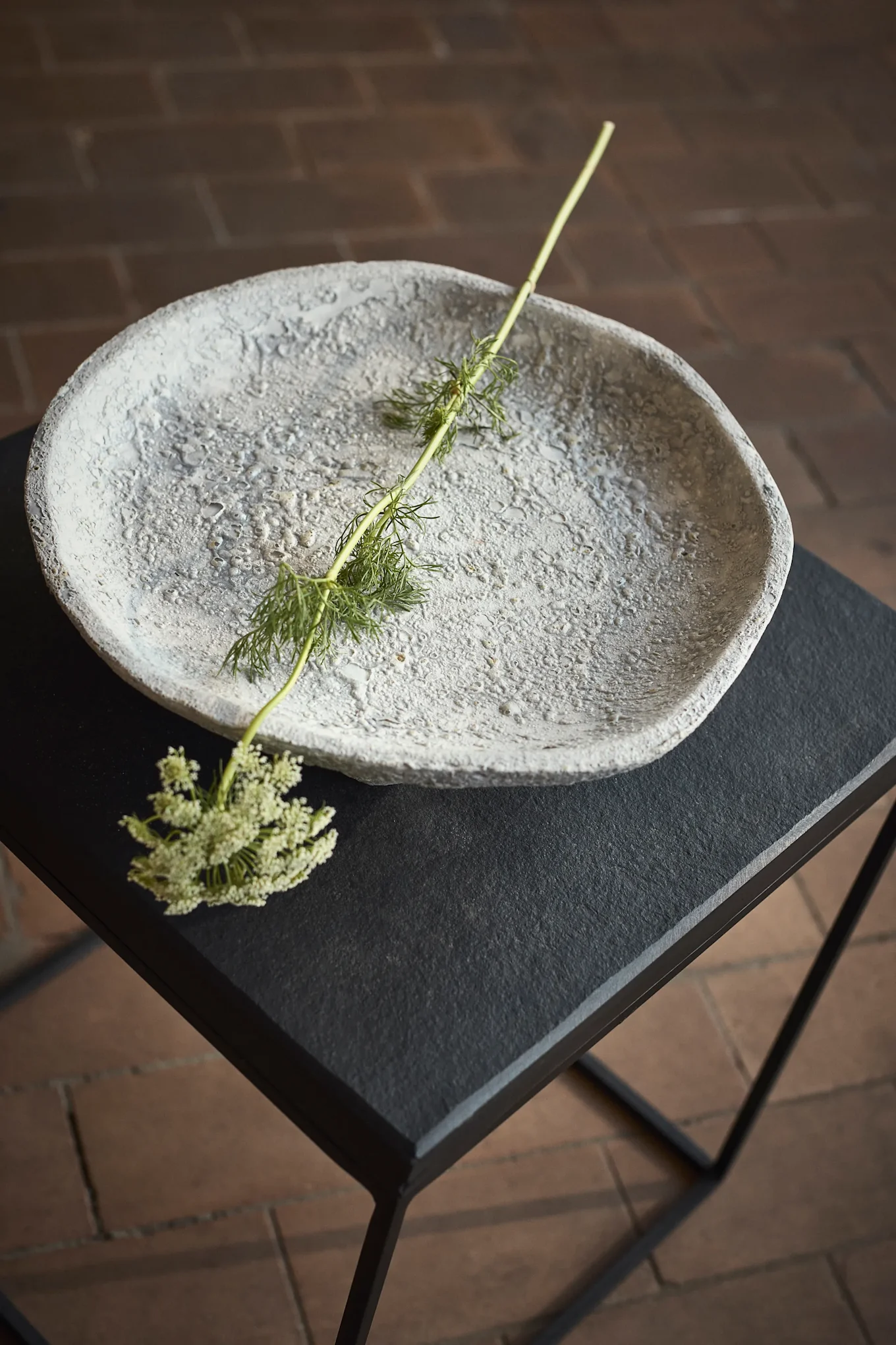 Bol gris texturé posé sur une table noire, orné d'une tige verte et de petites fleurs blanches.