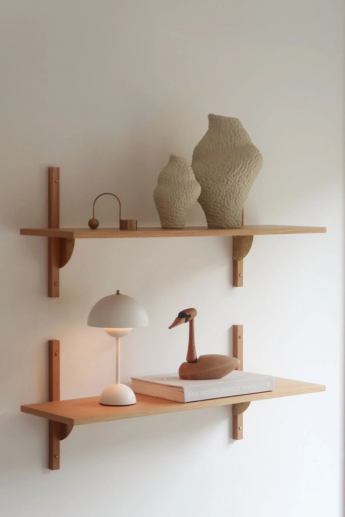 Deux étagères murales en bois clair avec de la décoration. L'étagère supérieure contient deux vases texturés et des objets en métal. L'étagère inférieure a une lampe blanche, un cygne en bois et un livre.