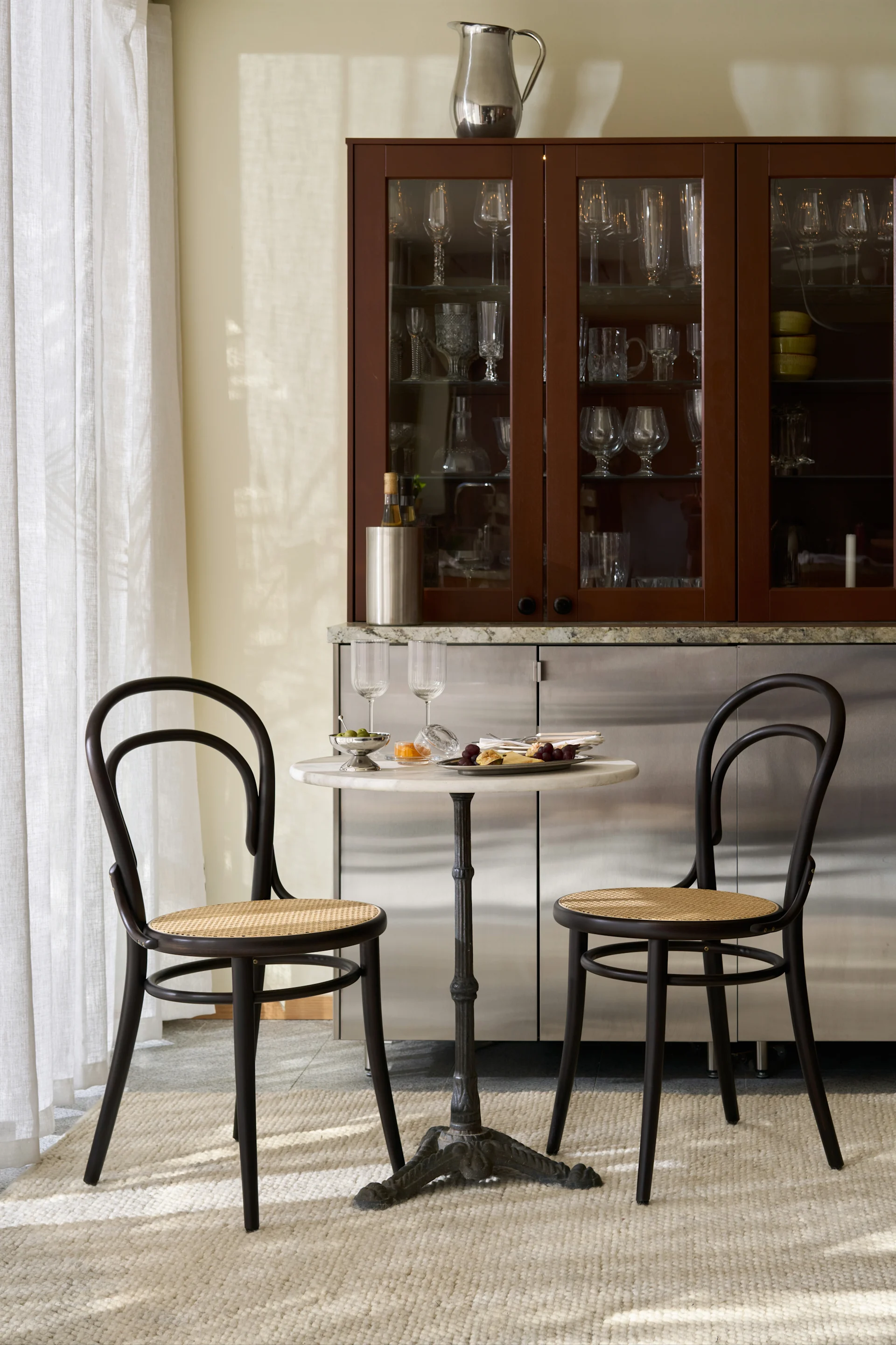 Un coin repas élégant avec une table de bistrot en marbre, deux chaises en bois foncé et un meuble bar rempli de verres.