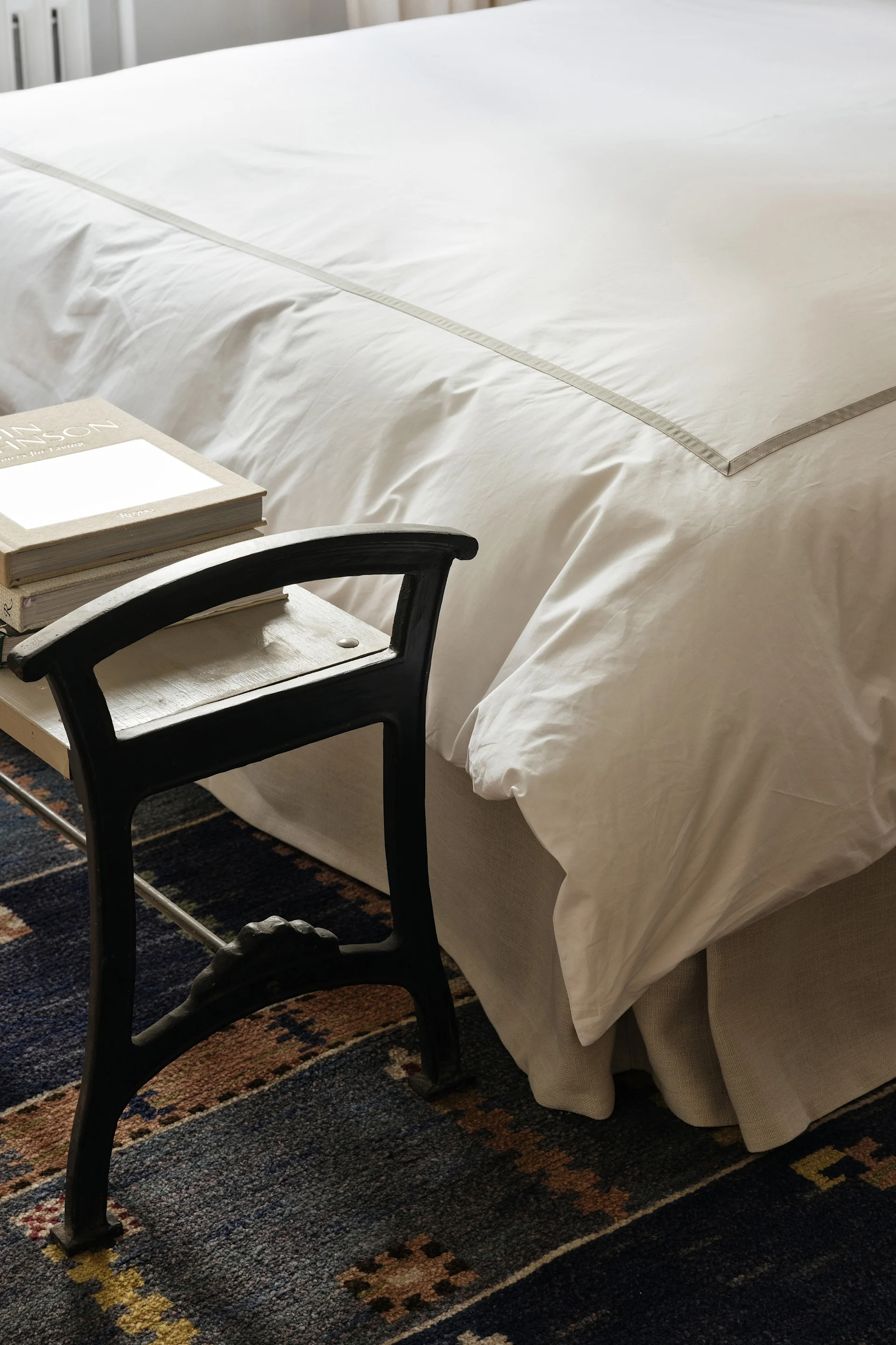 Un lit avec une literie blanche et une rayure grise à côté d'un tabouret sombre avec des livres sur un tapis bleu à motifs.