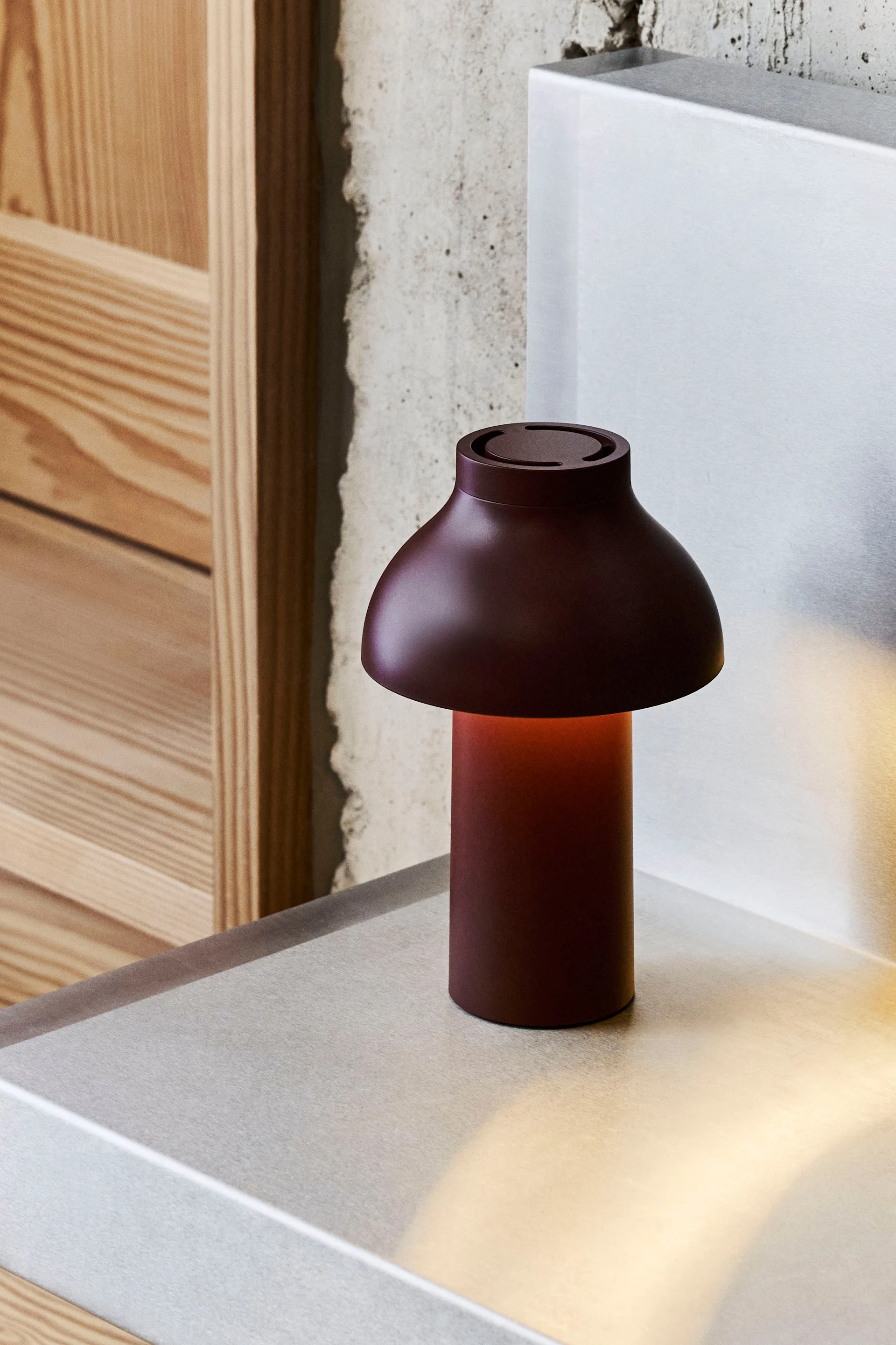Une lampe de table rouge foncé en forme de champignon avec une lueur chaude, posée sur une surface métallique, avec du béton et du bois en arrière-plan.