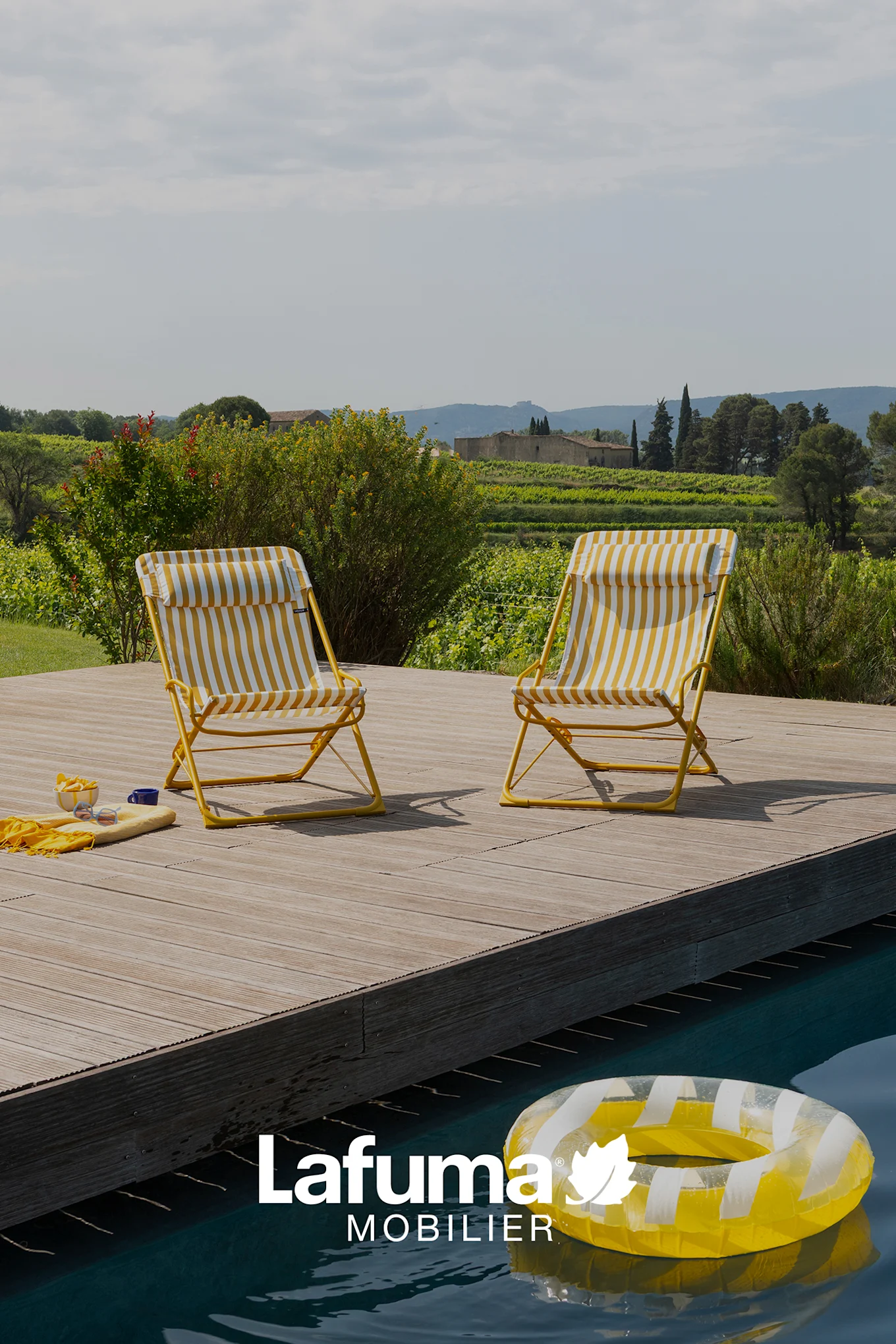 Deux chaises pliantes rayées jaunes et blanches au bord d'une piscine, avec un vignoble et des bâtiments en arrière-plan.