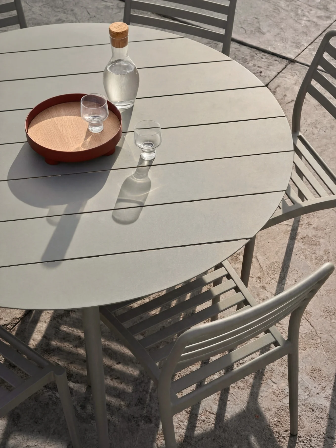 Un ensemble repas d'extérieur gris clair avec une table ronde à lattes et des chaises. Sur la table, une carafe d'eau et des verres sur un plateau.