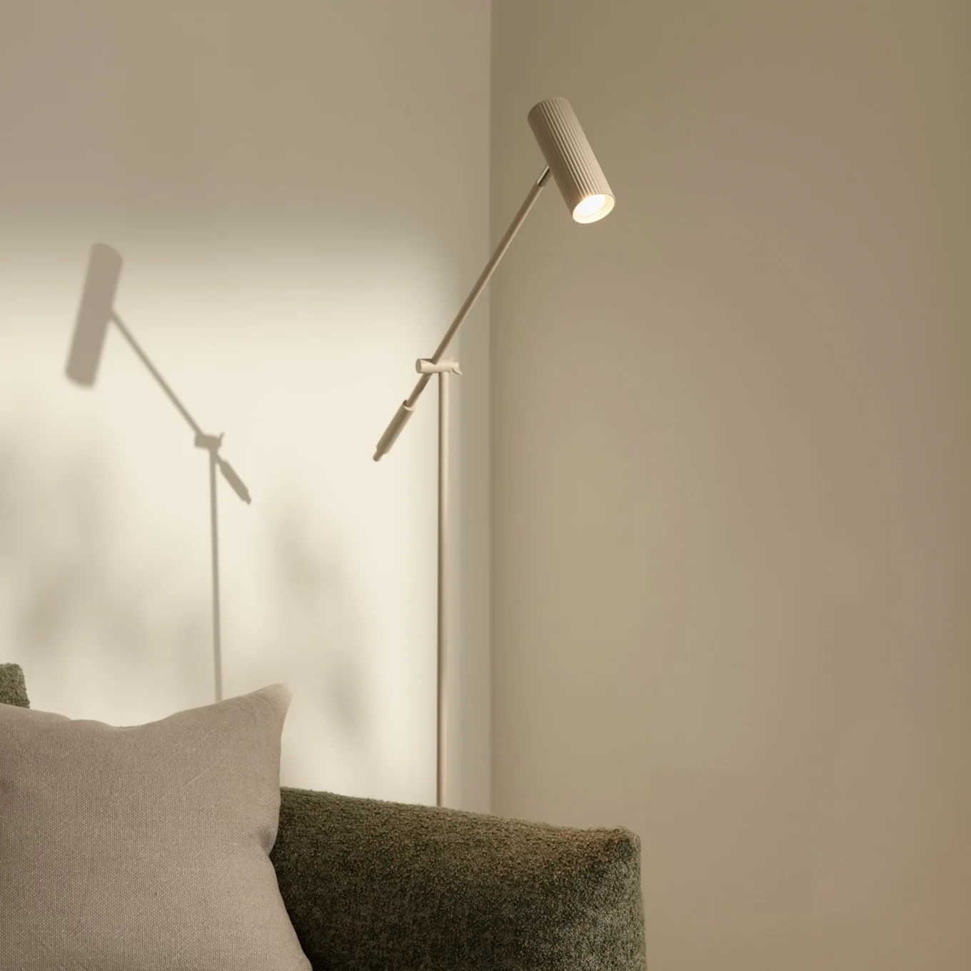 Un lampadaire moderne et élancé avec une tête côtelée éclairant un mur clair, à côté d'un canapé texturé vert avec un oreiller beige.