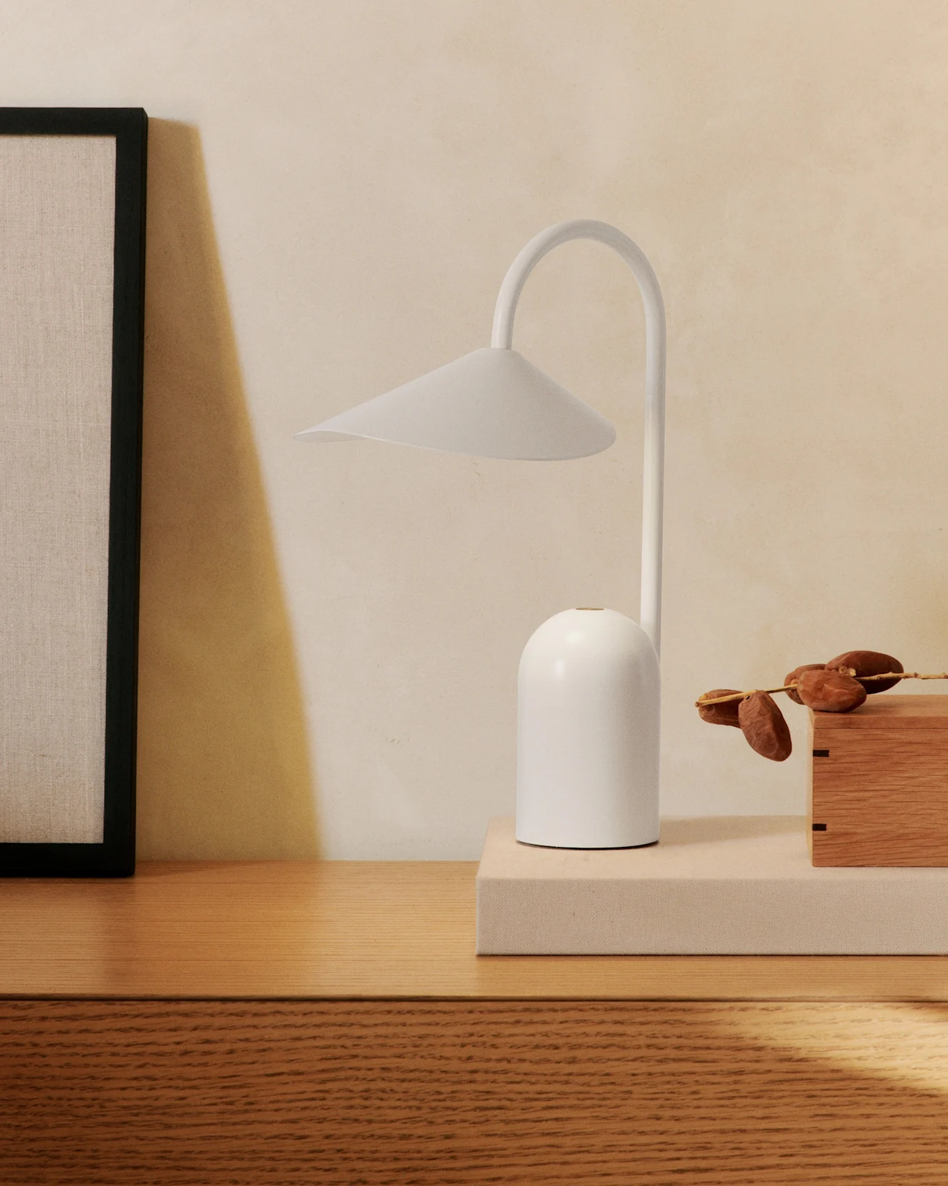 Une lampe de table blanche minimaliste avec un bras incurvé sur une commode en bois, à côté d'un tableau encadré et d'une boîte en bois avec des dattes.