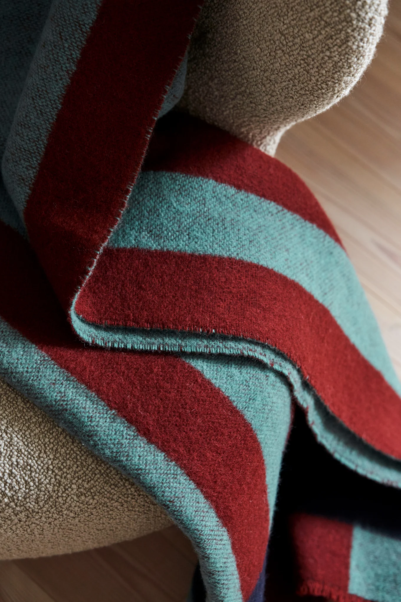 Couverture rayée pliée avec des bandes rouge foncé et sarcelle drapée sur un fauteuil texturé brun clair.