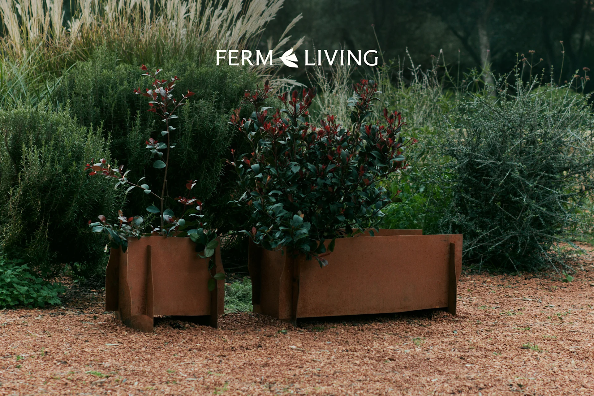 Deux jardinières métalliques patinées avec des plantes vertes et rouges sur gravier dans un jardin. Logo Ferm Living.